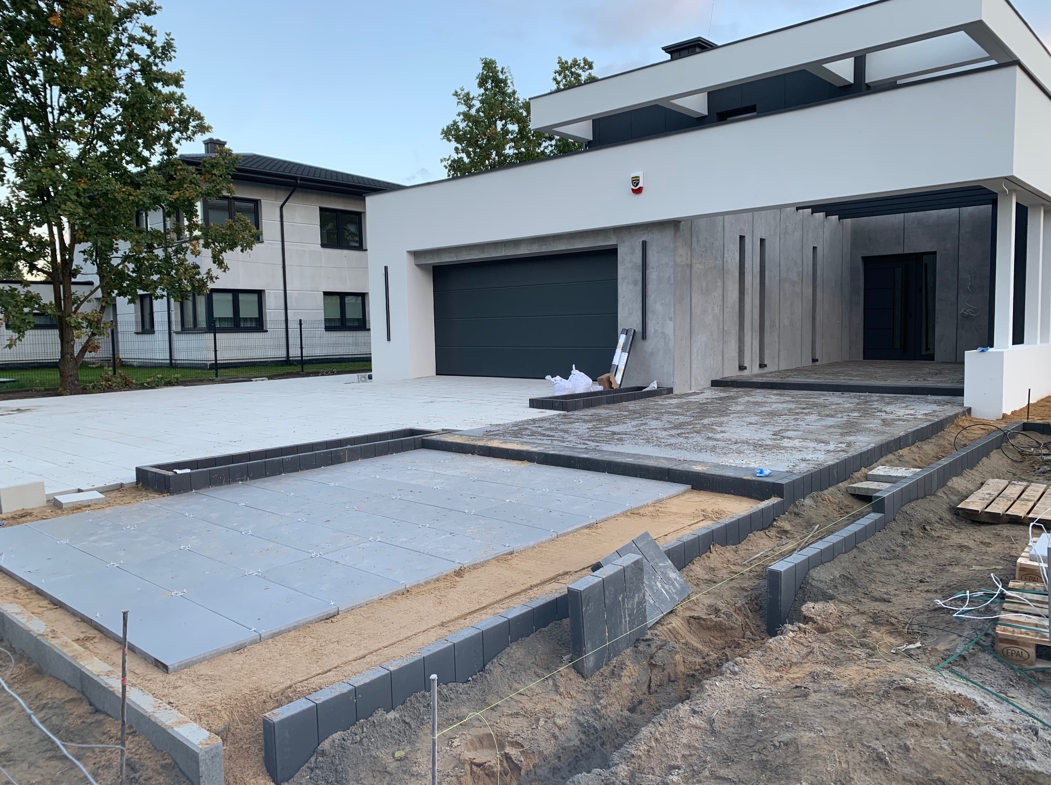 Etap prac przy układaniu szarej kostki brukowej na podsypce piaskowej, widoczne obrzeża z kostki, nowoczesny dom z garażem w tle.