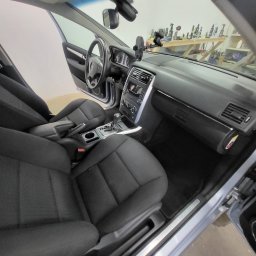 CarCarePoint_Detaling Samochodowy - Wnętrze samochodu przed praniem tapicerki: czarne fotele z materiału, deska rozdzielcza i kierownica. Na desce rozdzielczej uchwyt na telefon. W tle narzędzia.