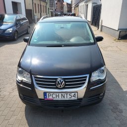 CarCarePoint_Detaling Samochodowy - Czarny Volkswagen na brukowanej ulicy, widok z przodu, przed korektą lakieru. Widoczne budynki w tle i odbicia na karoserii.