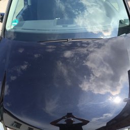 CarCarePoint_Detaling Samochodowy - Błyszcząca, czarna maska samochodu odbija chmury i niebo po korekcie lakieru. Widoczne odbicie osoby robiącej zdjęcie. Chromowany grill z logo VW na dole.