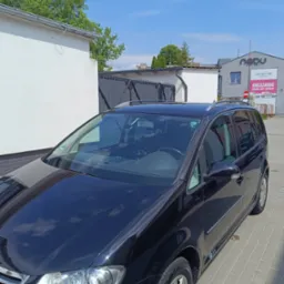 Błyszczący, czarny minivan po korekcie lakieru, zaparkowany na kostce brukowej przed budynkiem z reklamą oklejania pojazdów. Słoneczny dzień.