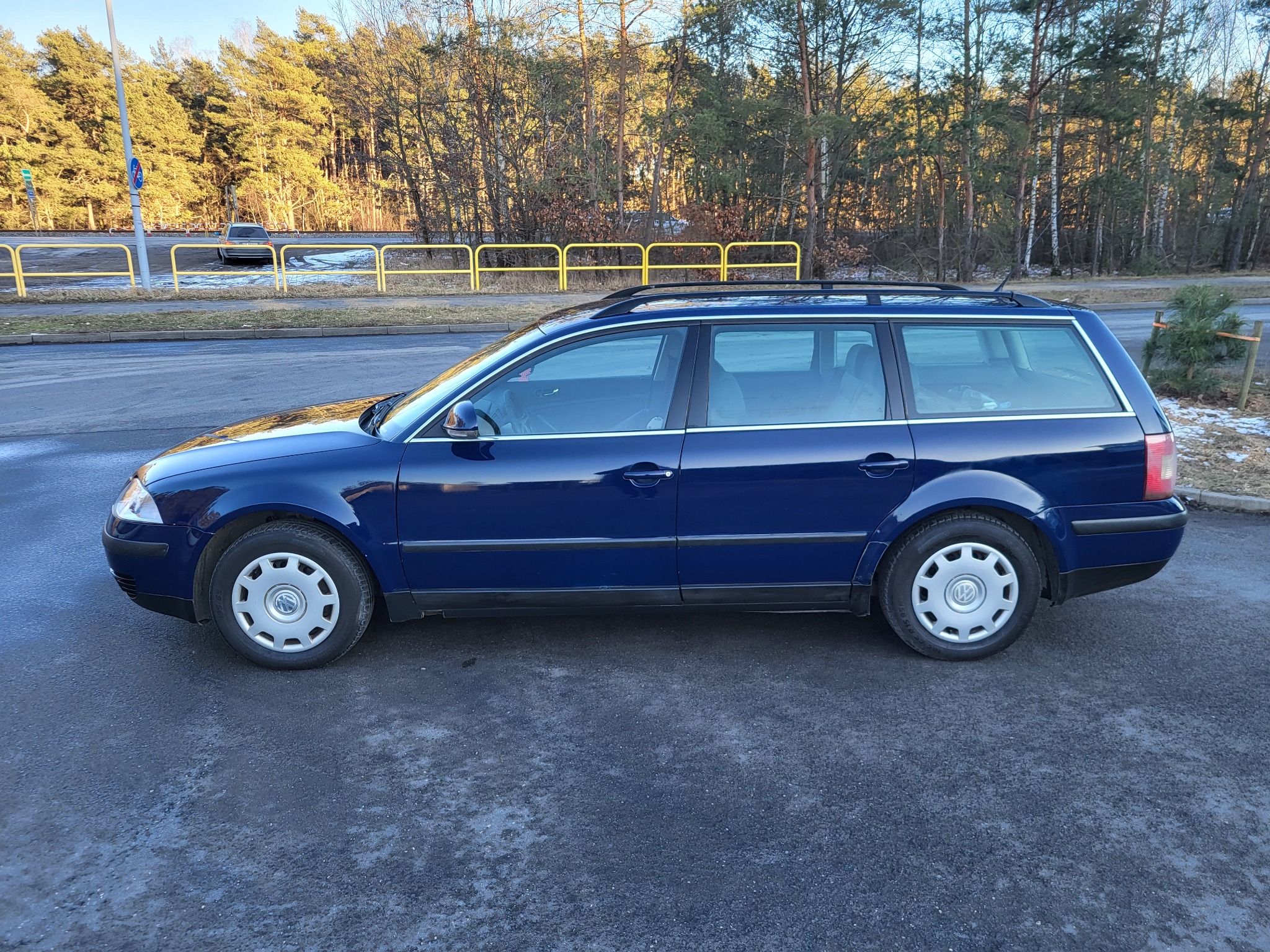 Błyszczący, granatowy VW Passat Variant po korekcie lakieru, zaparkowany na asfalcie, z widocznym otoczeniem drzew i żółtymi barierkami. Słoneczny dzień.