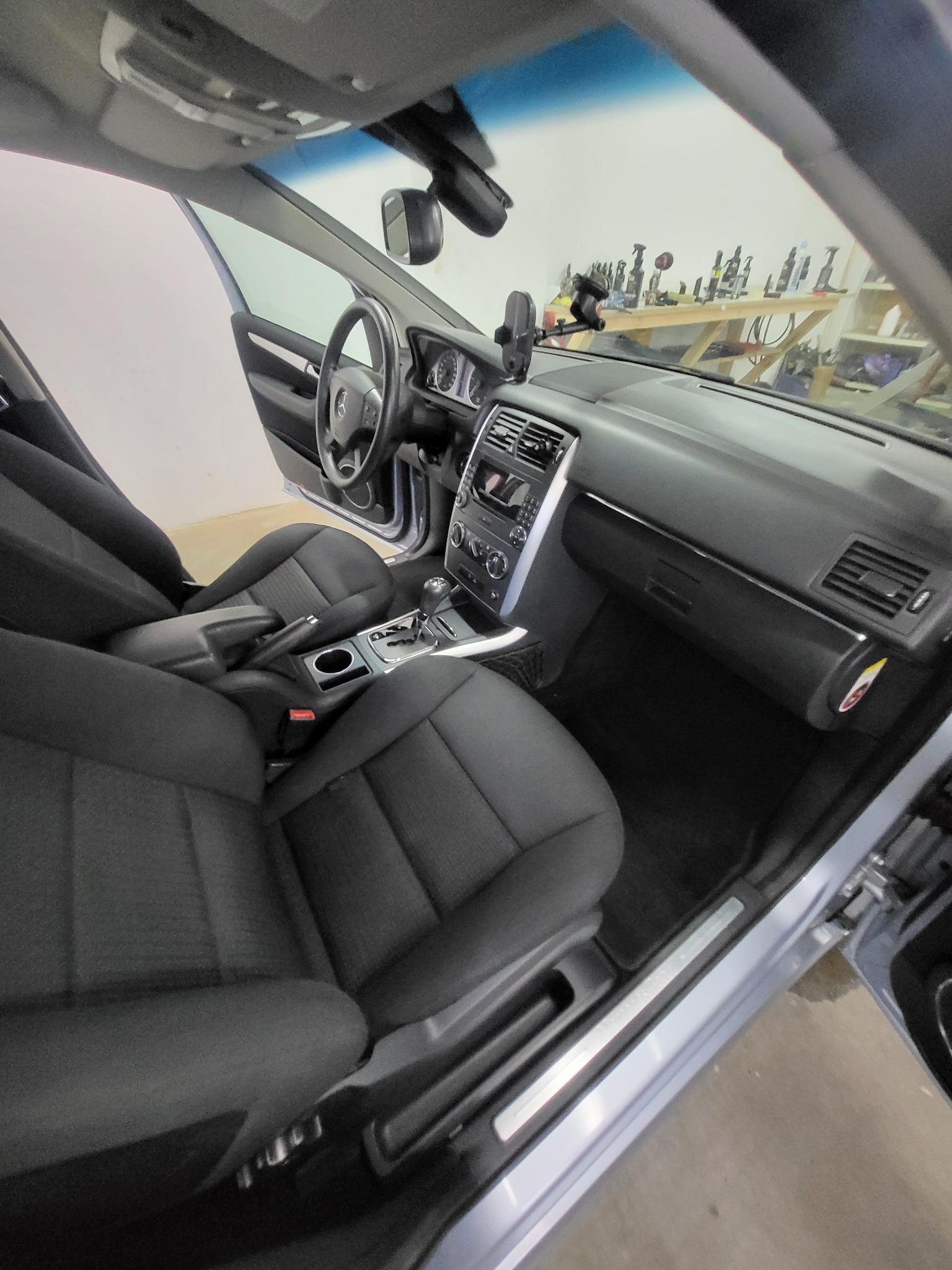 Wnętrze samochodu przed praniem tapicerki: czarne fotele z materiału, deska rozdzielcza i kierownica. Na desce rozdzielczej uchwyt na telefon. W tle narzędzia.