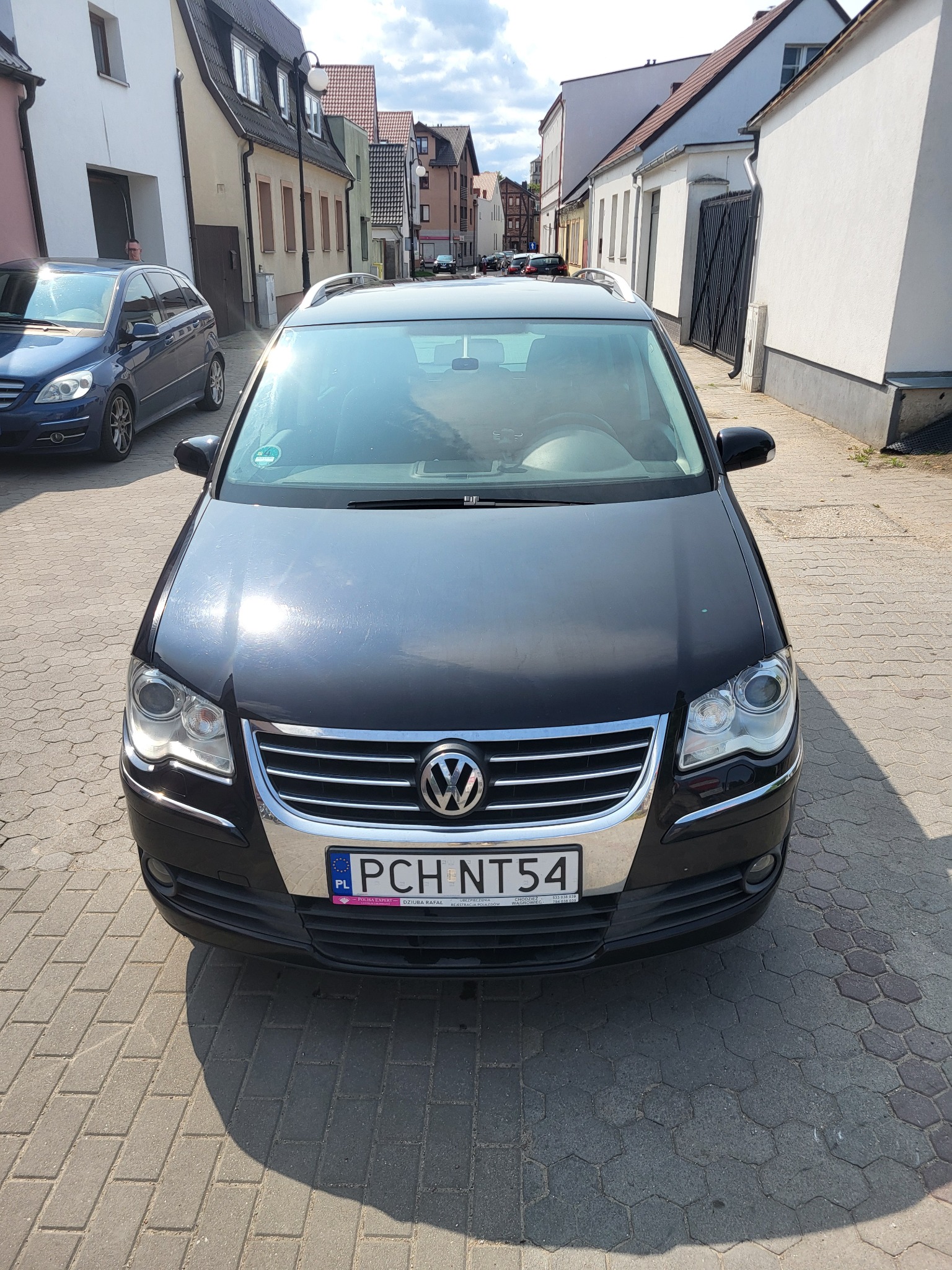 Czarny Volkswagen na brukowanej ulicy, widok z przodu, przed korektą lakieru. Widoczne budynki w tle i odbicia na karoserii.