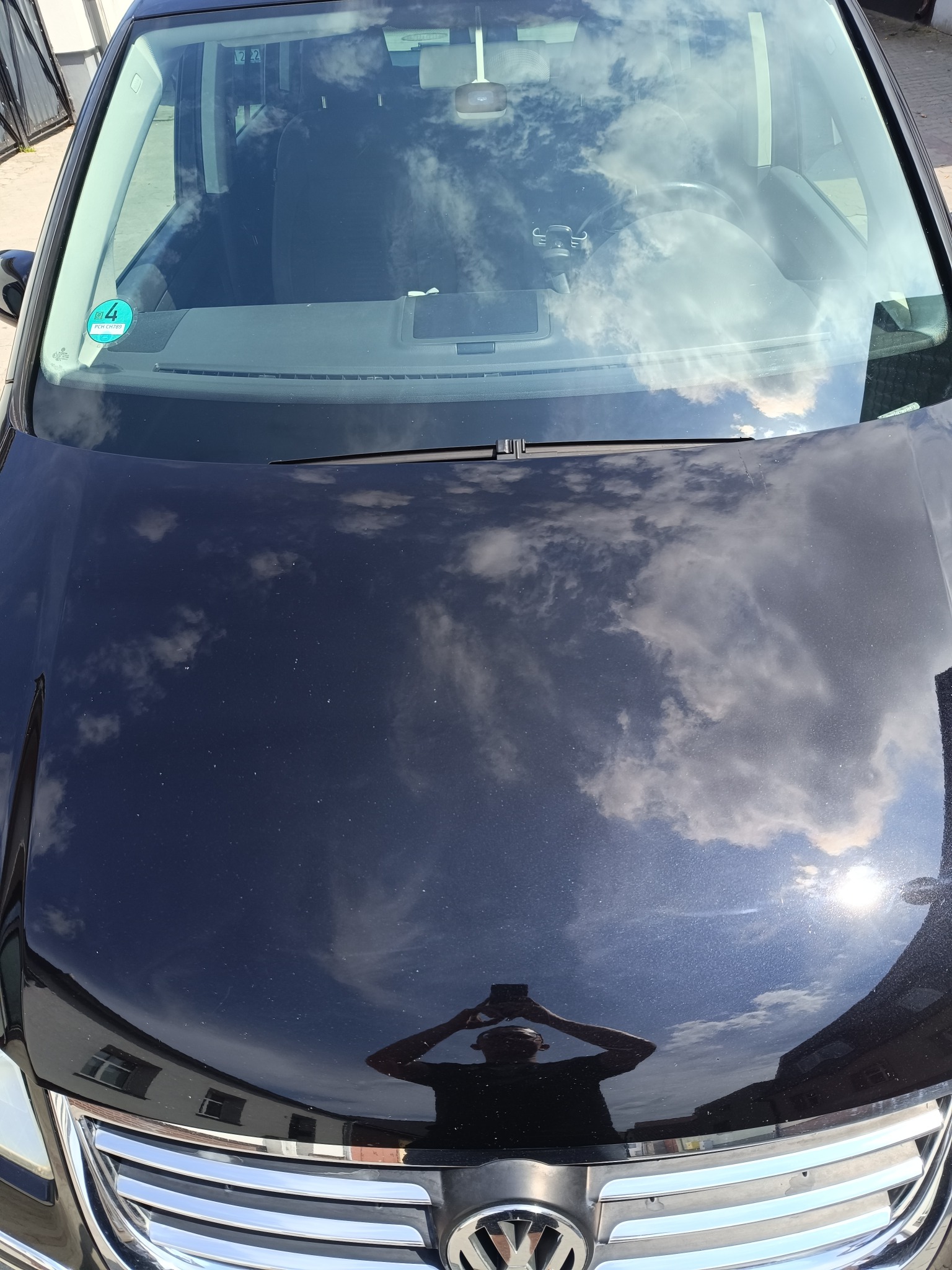 Błyszcząca, czarna maska samochodu odbija chmury i niebo po korekcie lakieru. Widoczne odbicie osoby robiącej zdjęcie. Chromowany grill z logo VW na dole.