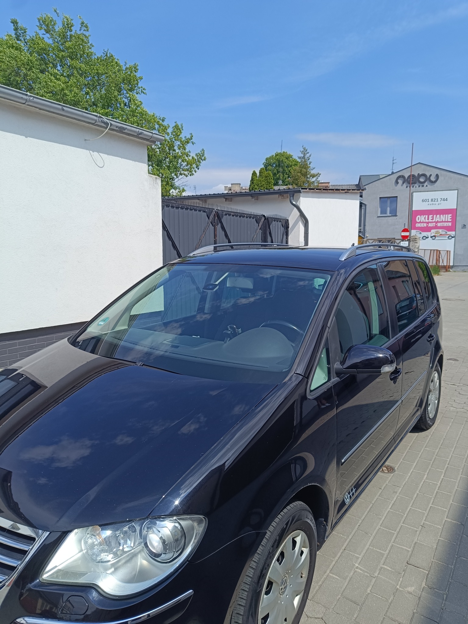 Błyszczący, czarny minivan po korekcie lakieru, zaparkowany na kostce brukowej przed budynkiem z reklamą oklejania pojazdów. Słoneczny dzień.