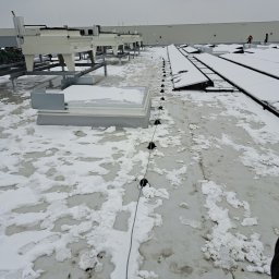 Denys Syrota - Instalacja odgromowa na płaskim dachu pokrytym śniegiem, widoczne elementy mocujące i przewody, w tle urządzenia chłodnicze i pracownicy w odzieży roboczej.