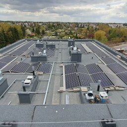 Denys Syrota - Widok z góry na instalację fotowoltaiczną na płaskim dachu budynku, widoczne panele, system mocowania, elementy wentylacyjne i fragment panoramy miasta w tle.