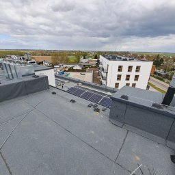 Denys Syrota - Widok z dachu budynku wielorodzinnego na miasto, widoczne panele słoneczne, instalacja odgromowa w trakcie montażu, elementy wentylacji i kominy.