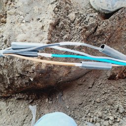 Denys Syrota - Połączenie kabli elektrycznych w ziemi, widoczne izolowane przewody o różnych kolorach, złączki termokurczliwe i fragment buta roboczego w tle.