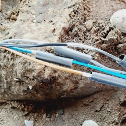 Denys Syrota - Kable elektryczne w wykopie ziemnym, zabezpieczone czarnymi rurkami termokurczliwymi, widoczne fragmenty butów roboczych i narzędzia.