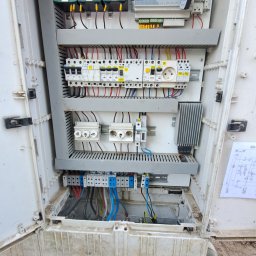 Denys Syrota - Otwarta, brudna szafa elektryczna z widocznymi bezpiecznikami, przewodami i schematem na drzwiach, sfotografowana z góry z widocznymi butami osoby stojącej przed nią.