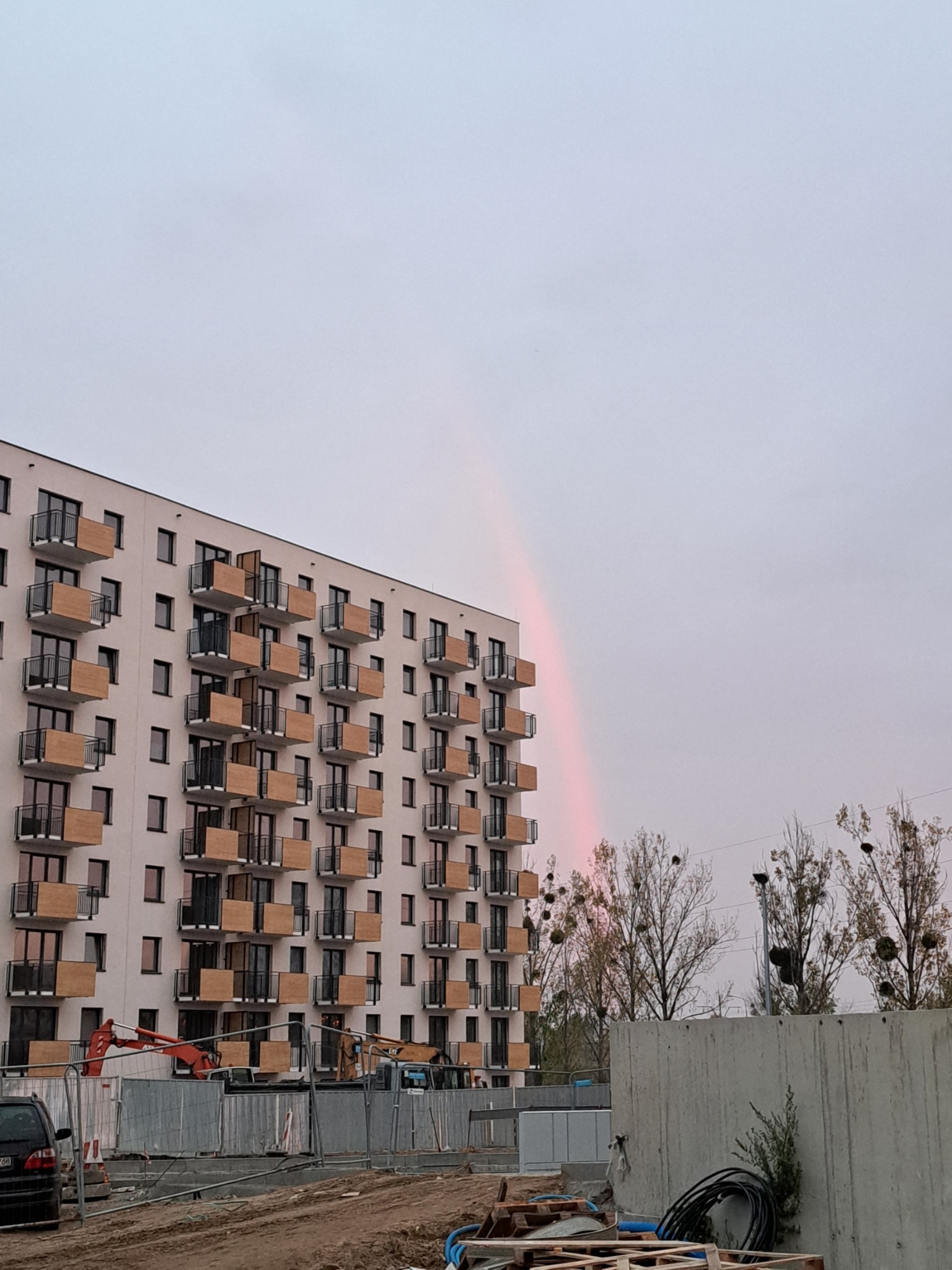 Nowy budynek wielorodzinny z balkonami w trakcie budowy, widoczny tęczowy łuk na pochmurnym niebie, maszyny budowlane i tymczasowe ogrodzenie.