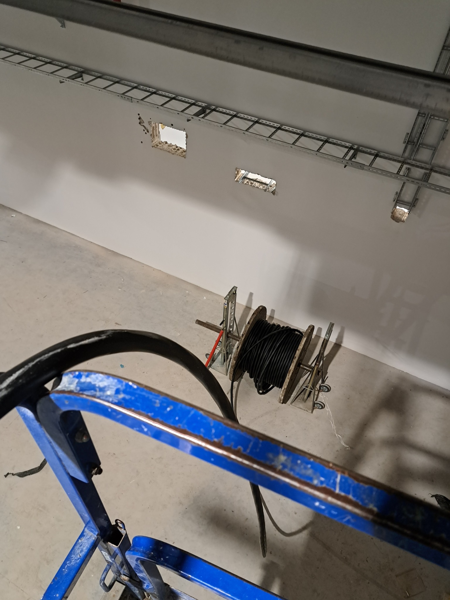 Widok z góry na szpulę z czarnym kablem elektrycznym na metalowym stojaku z kółkami, umieszczoną na betonowej podłodze w pobliżu białej ściany z otworami i metalowymi korytkami kablowymi...