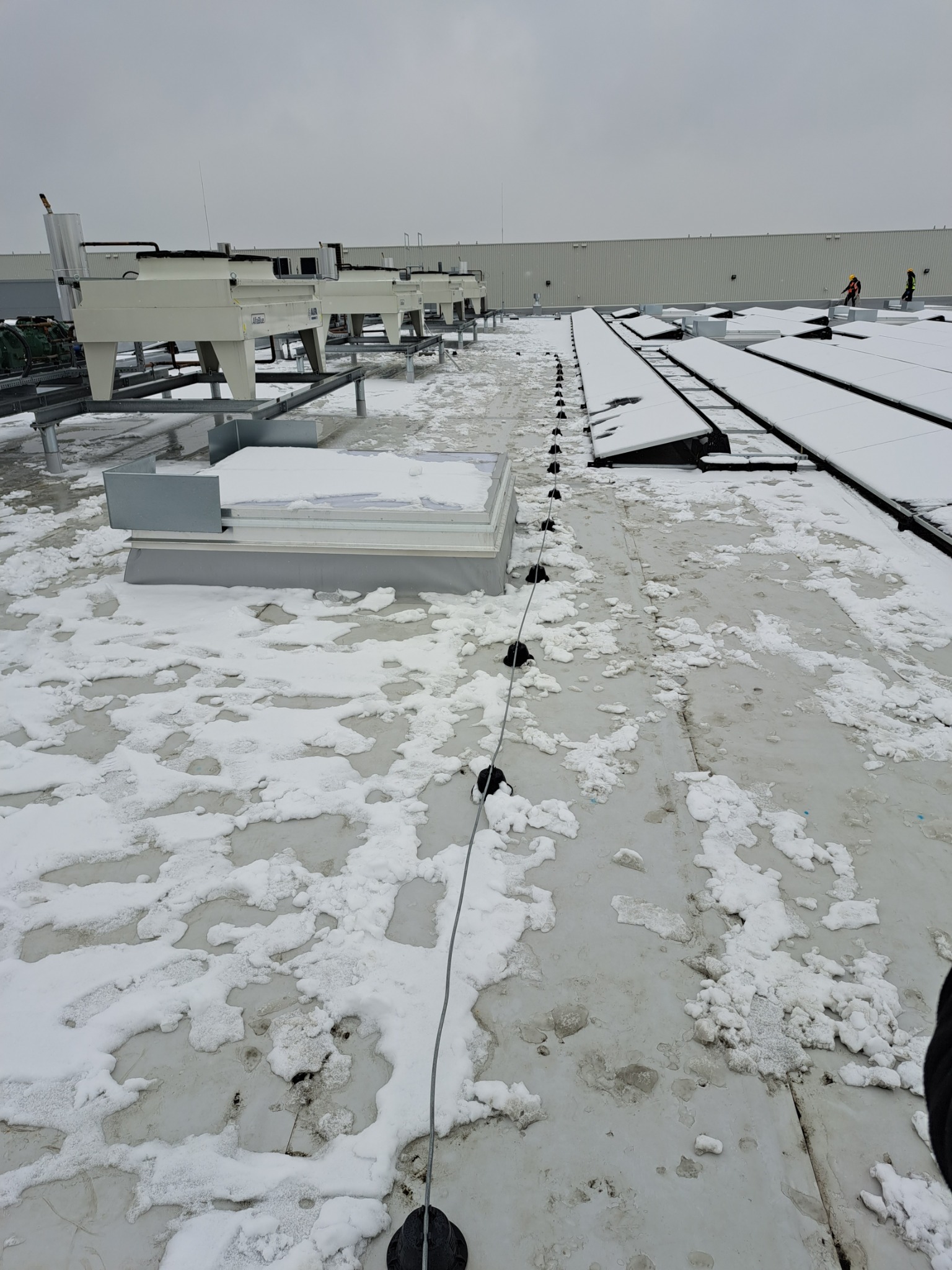 Instalacja odgromowa na płaskim dachu pokrytym śniegiem, widoczne elementy mocujące i przewody, w tle urządzenia chłodnicze i pracownicy w odzieży roboczej.