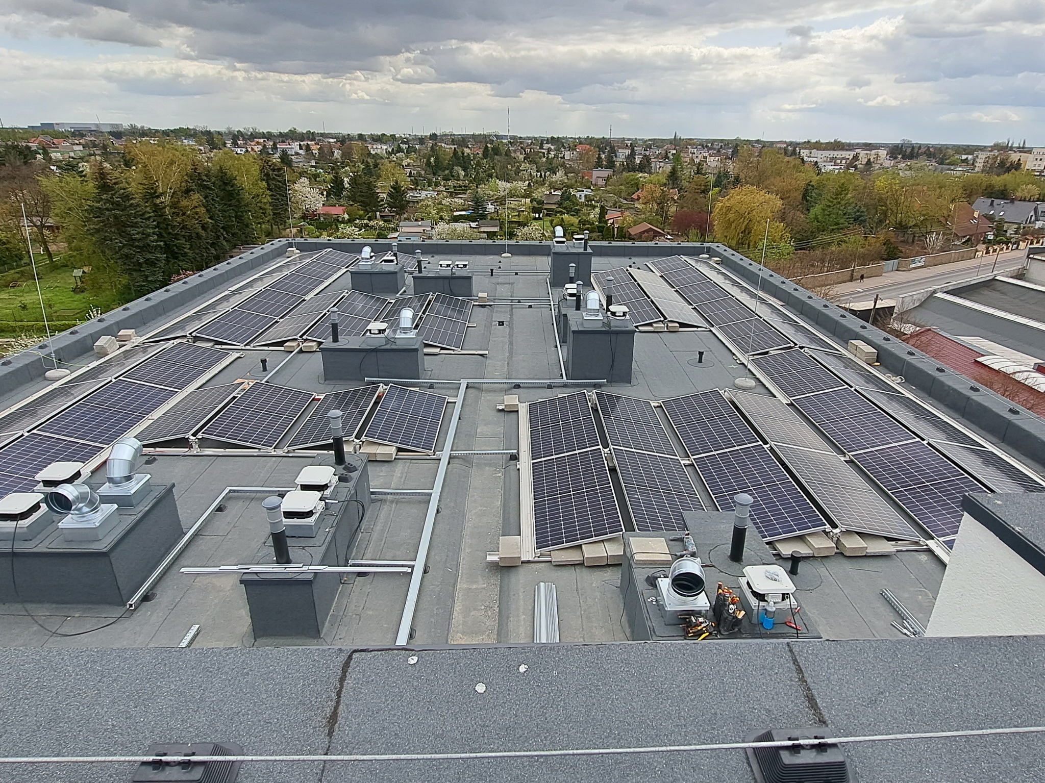 Widok z góry na instalację fotowoltaiczną na płaskim dachu budynku, widoczne panele, system mocowania, elementy wentylacyjne i fragment panoramy miasta w tle.