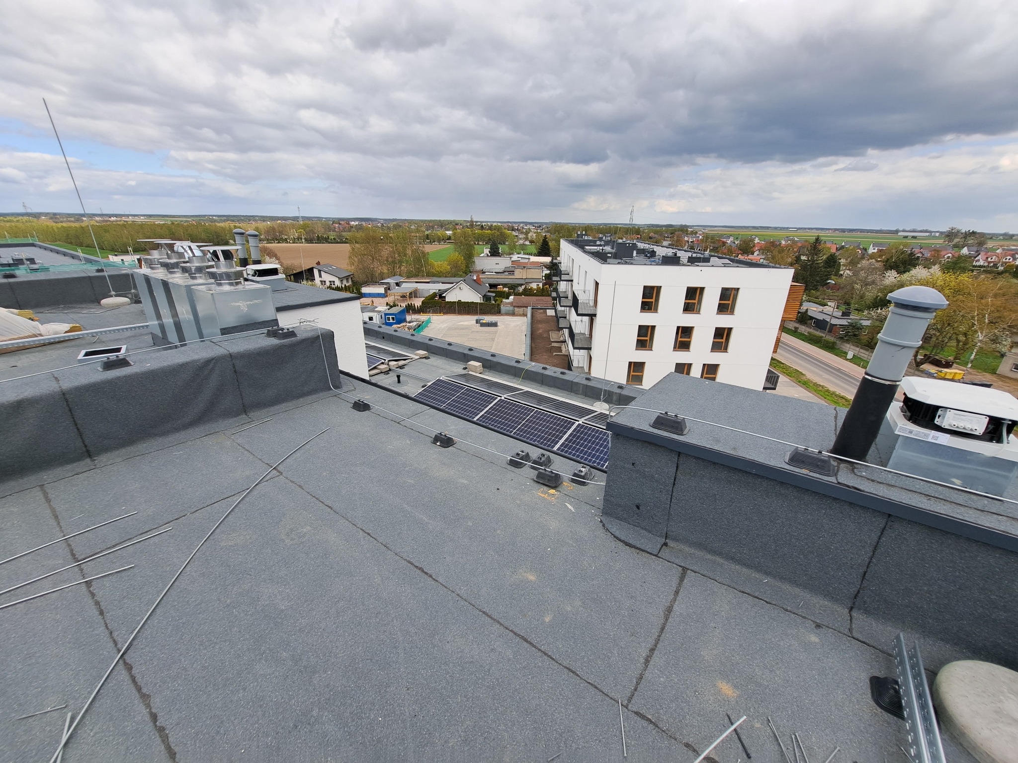 Widok z dachu budynku wielorodzinnego na miasto, widoczne panele słoneczne, instalacja odgromowa w trakcie montażu, elementy wentylacji i kominy.