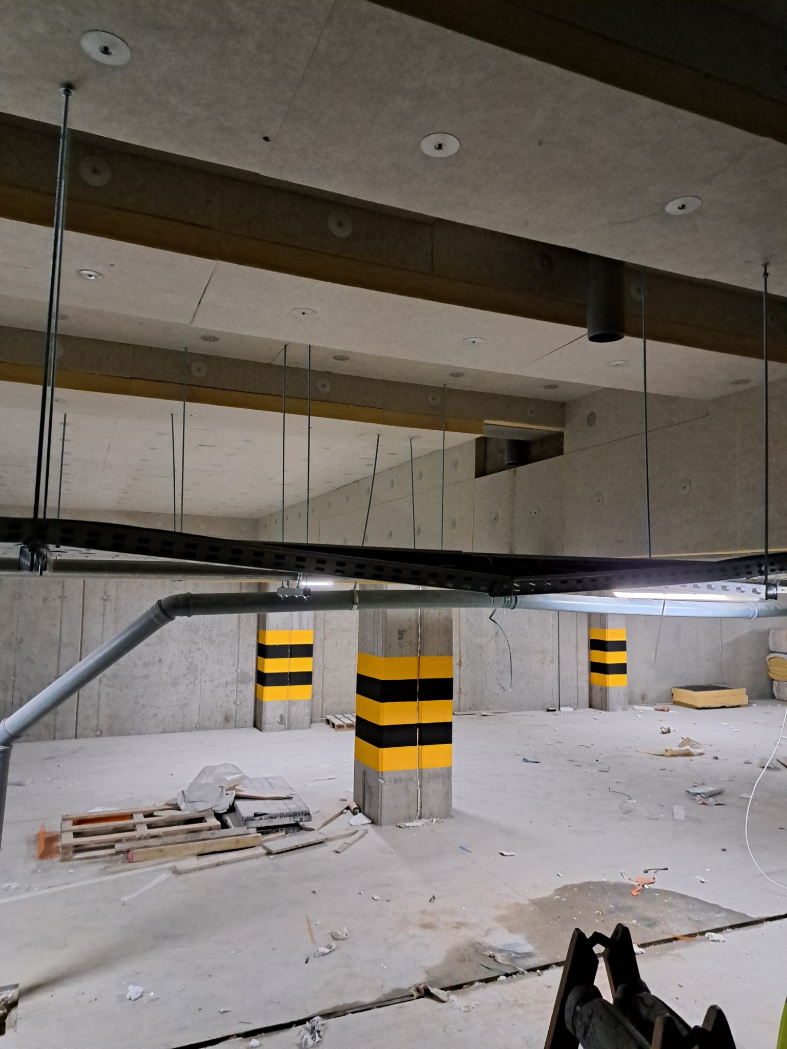 Wnętrze garażu podziemnego w trakcie prac instalacyjnych: widoczne koryta kablowe pod sufitem, rury wentylacyjne i słupy oznaczone pasami ostrzegawczymi.