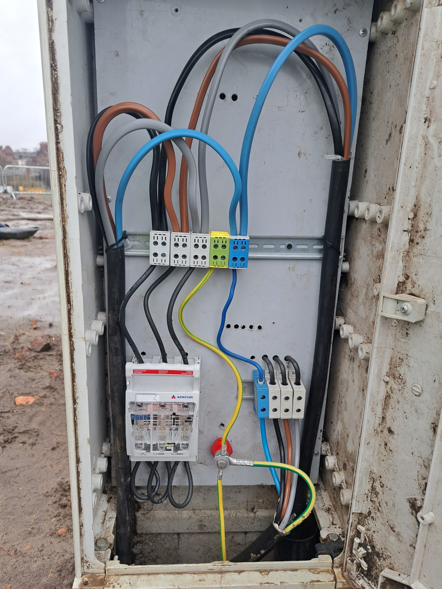 Otwarta skrzynka elektryczna z widocznym licznikiem, przewodami o różnych kolorach i złączkami, zamontowana na zewnątrz budynku, w tle teren budowy.