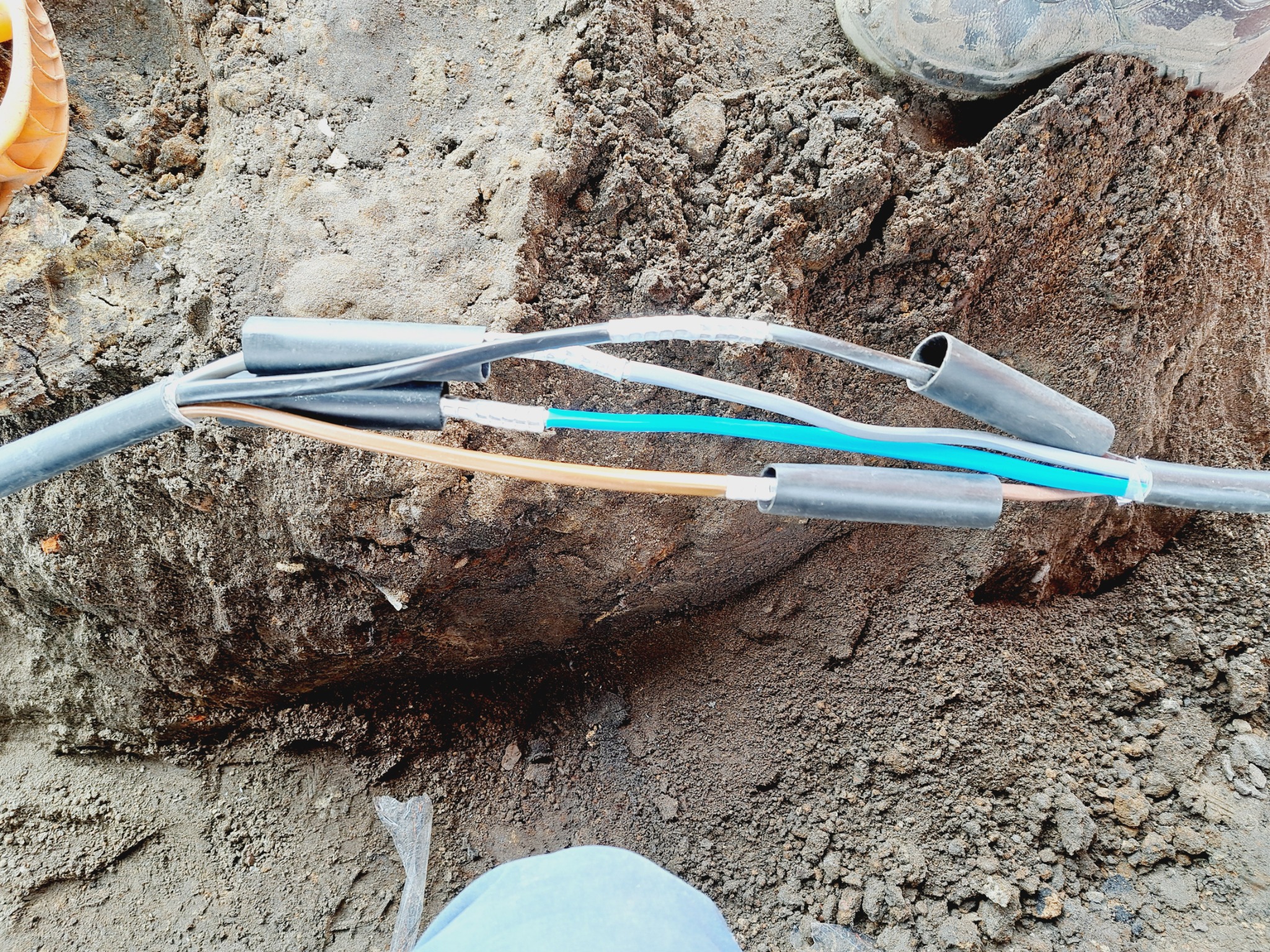 Połączenie kabli elektrycznych w ziemi, widoczne izolowane przewody o różnych kolorach, złączki termokurczliwe i fragment buta roboczego w tle.