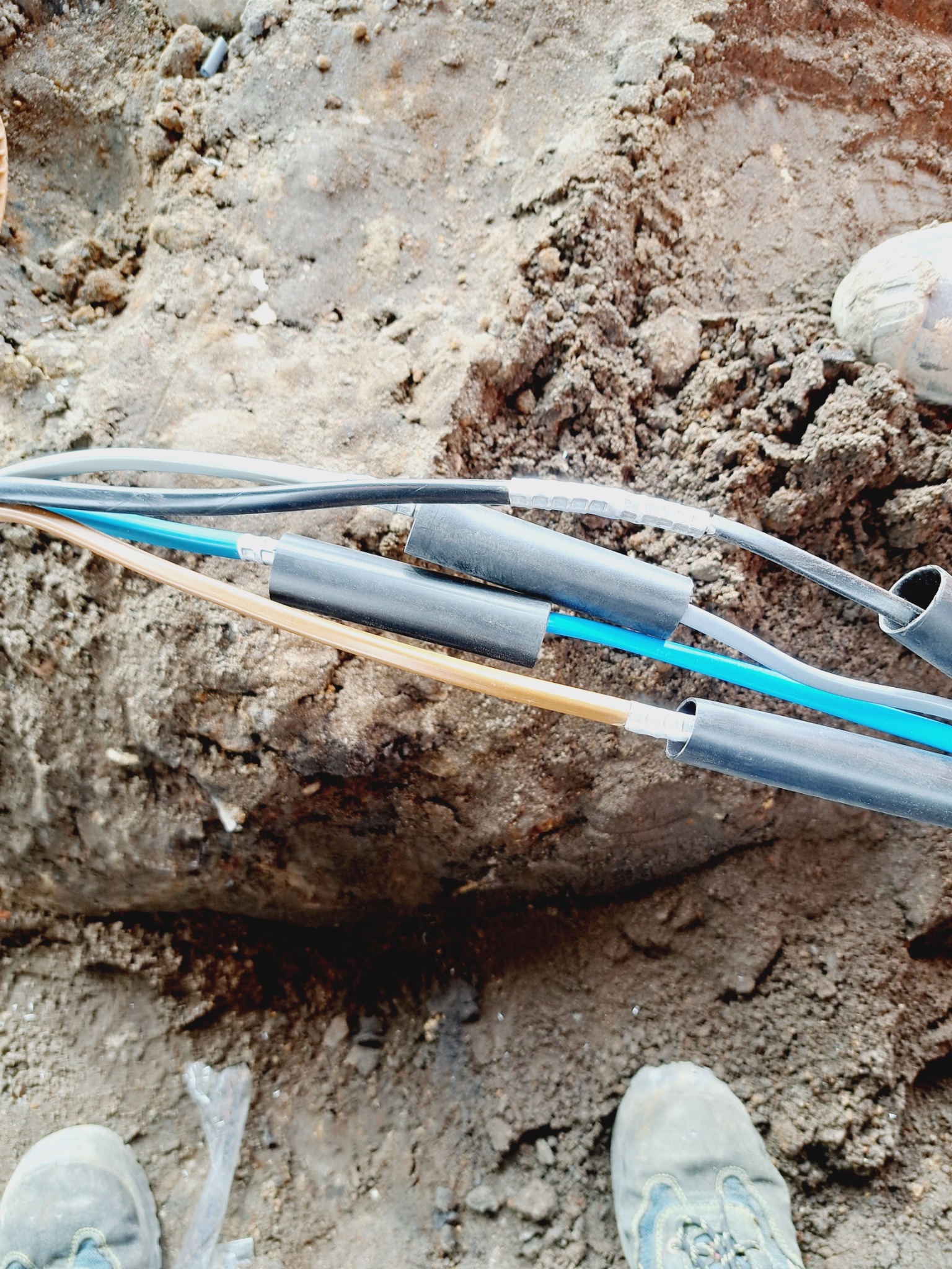 Kable elektryczne w wykopie ziemnym, zabezpieczone czarnymi rurkami termokurczliwymi, widoczne fragmenty butów roboczych i narzędzia.