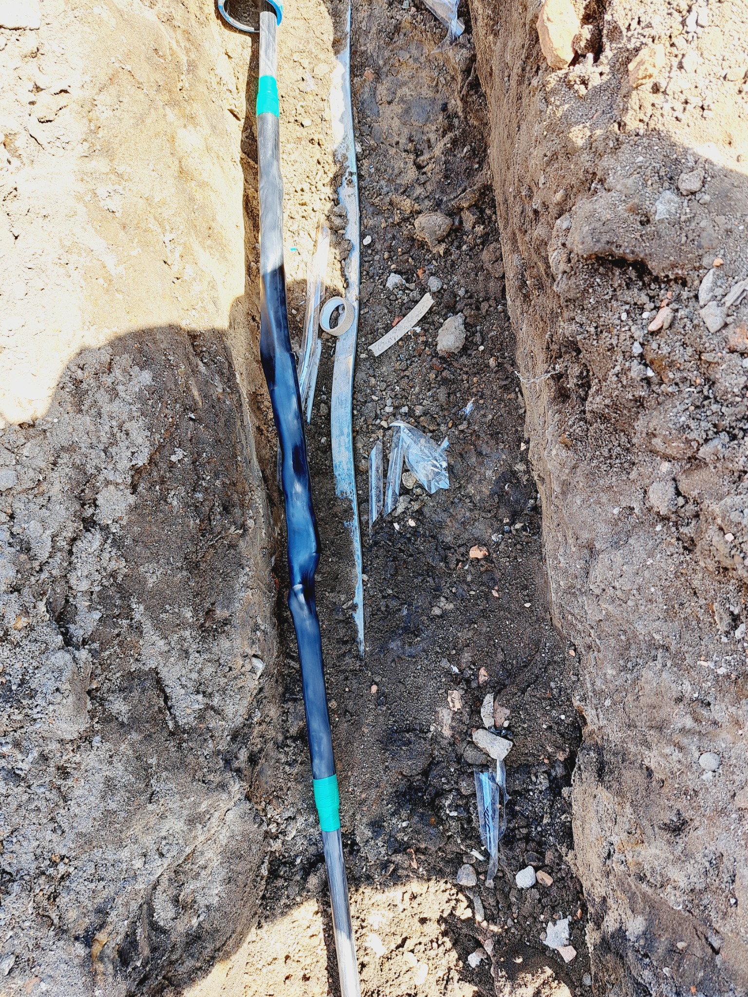 Wykop z ułożonym kablem elektrycznym w czarnej izolacji, zabezpieczonym taśmą, widoczne fragmenty gruzu i folii.