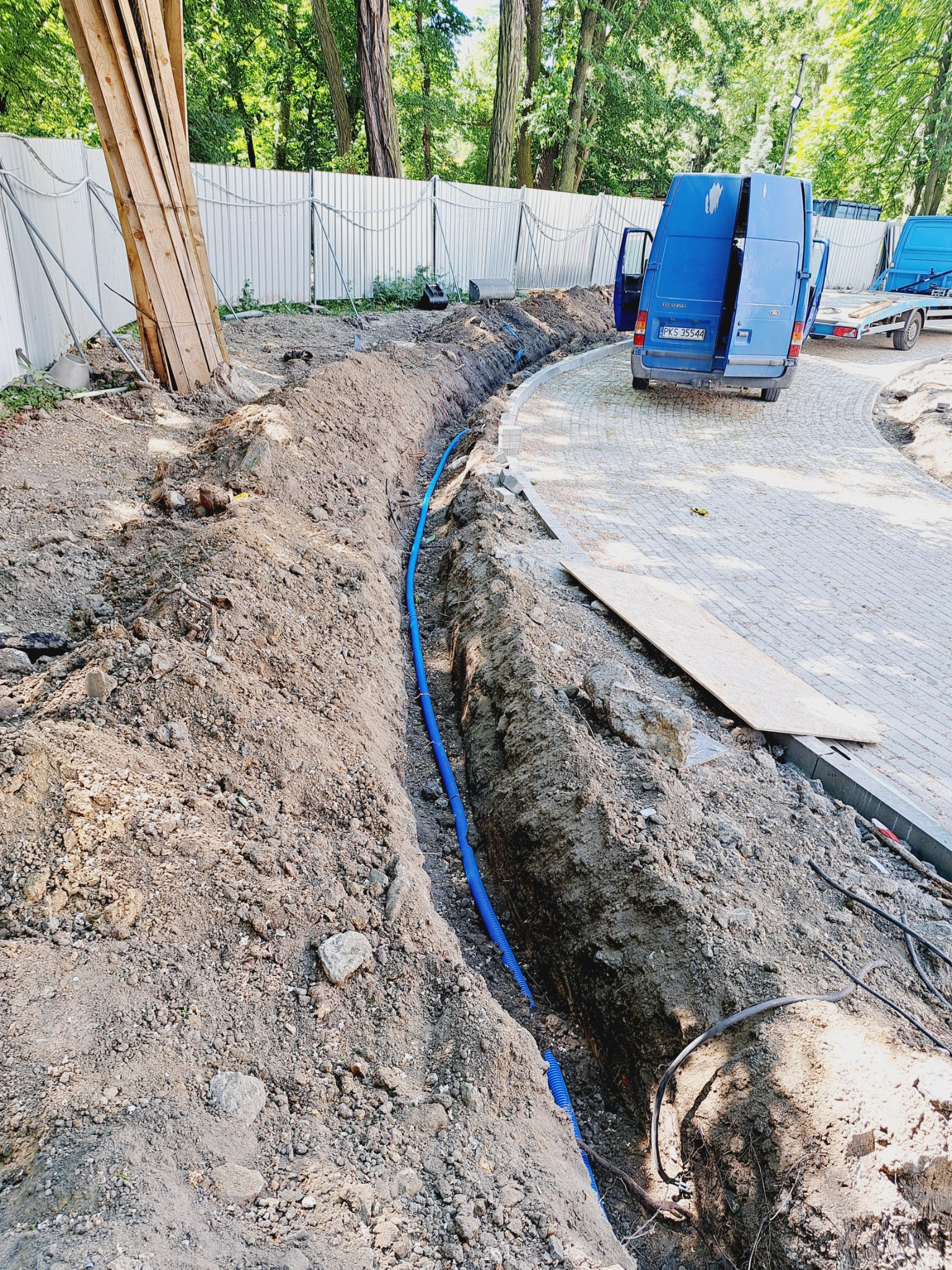 Wykop z ułożoną niebieską rurą karbowaną, prawdopodobnie peszel, widoczny niebieski bus z otwartymi drzwiami i przyczepką na kostce brukowej, teren budowy zabezpieczony płotem.