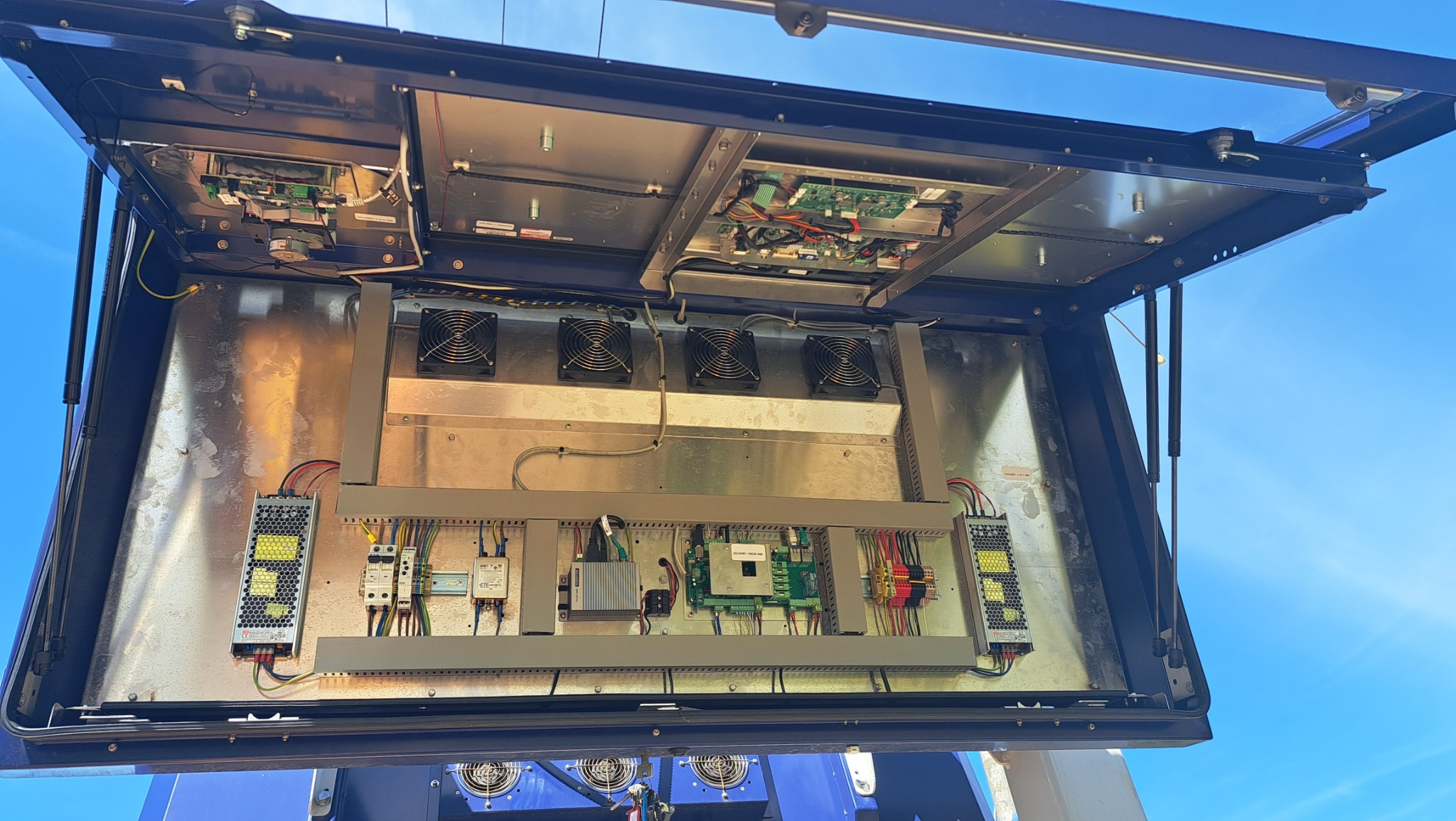 Otwarta szafa sterownicza z widocznymi elementami elektrycznymi: wentylatory, zasilacze, bezpieczniki i płyty PCB, zamontowana na zewnątrz, na tle błękitnego nieba.