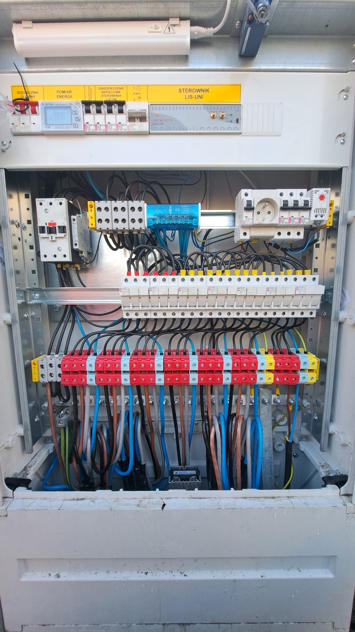 Otwarta skrzynka elektryczna z widocznymi bezpiecznikami, sterownikiem LIS-UNI, pomiarem energii, okablowaniem w kolorach niebieskim, czarnym, brązowym i zielono-żółtym, zamontowana na szarej...