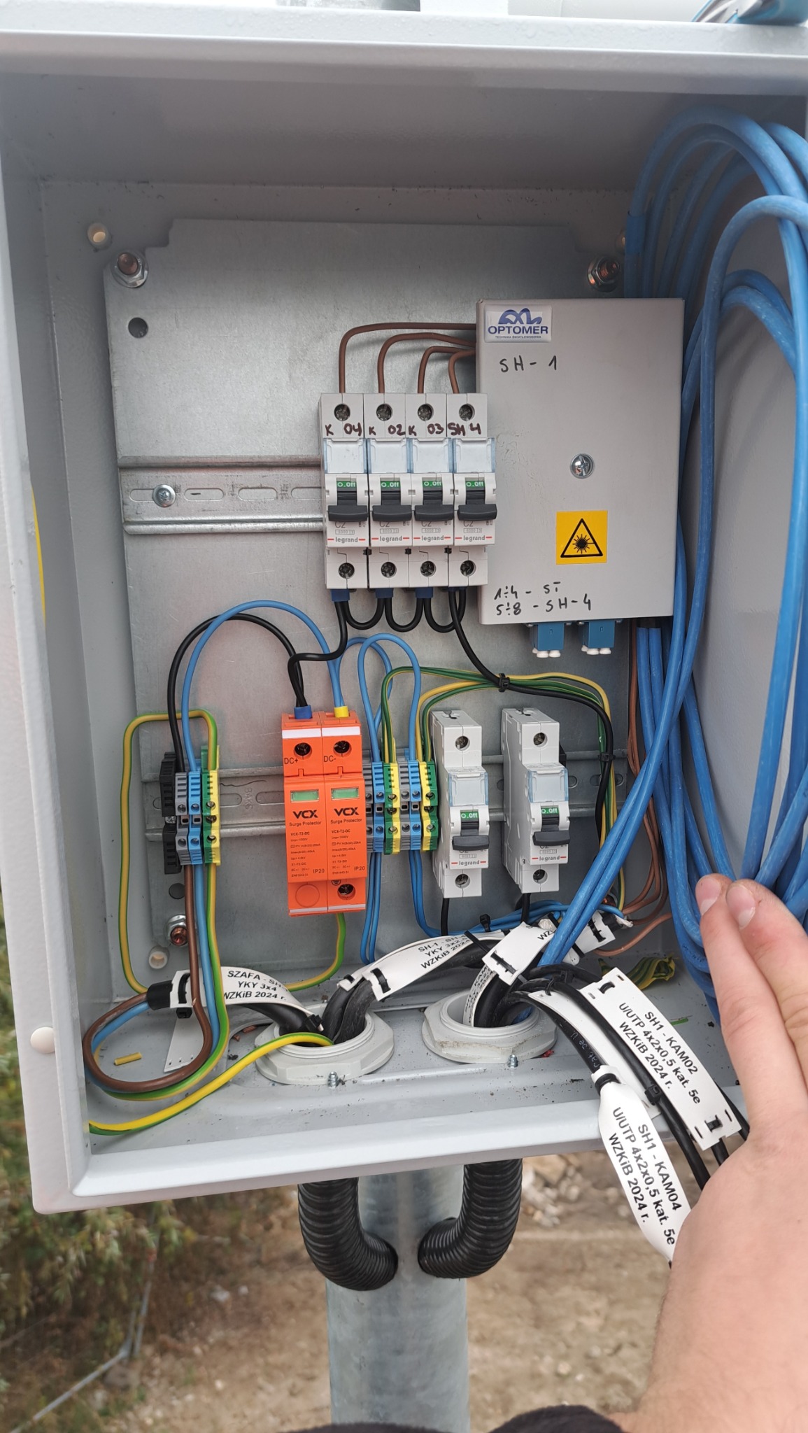 Otwarta skrzynka elektryczna na słupie z widocznymi zabezpieczeniami przepięciowymi, wyłącznikami, okablowaniem i oznaczeniami przewodów. Na białej obudowie skrzynki widoczny znak ostrzegawczy.