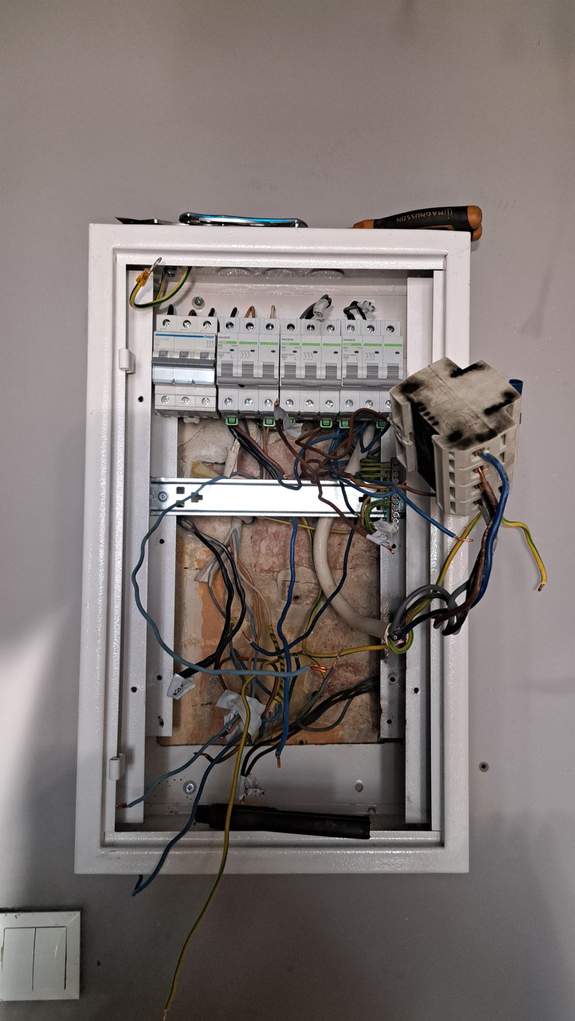 Otwarta skrzynka elektryczna z bezpiecznikami i przewodami w trakcie podłączania, widoczne narzędzia na górnej krawędzi skrzynki.