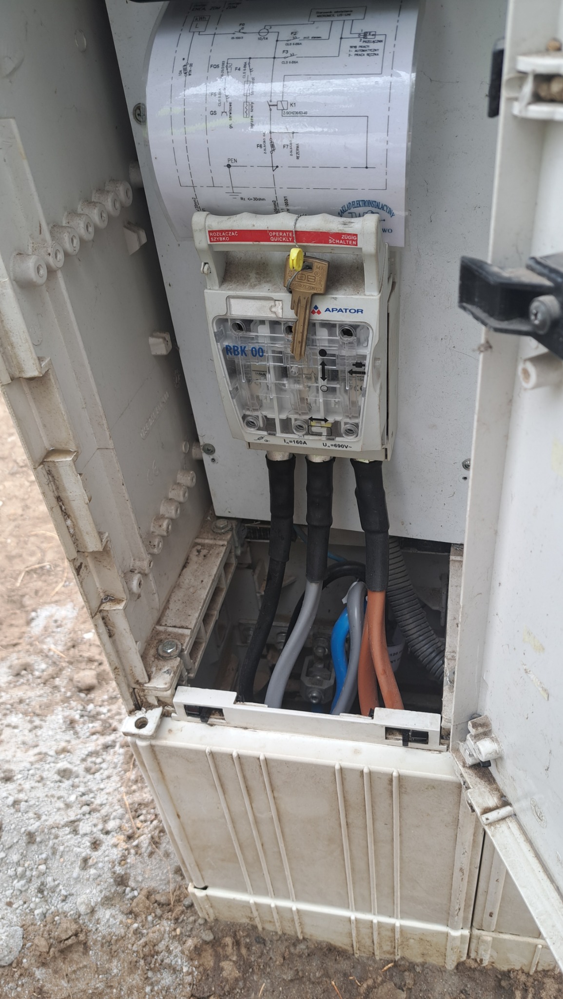 Otwarta skrzynka elektryczna z widocznym zabezpieczeniem RBK 00, przewodami w różnych kolorach i schematem instalacji przymocowanym do wewnętrznej strony pokrywy. Klucz w stacyjce.