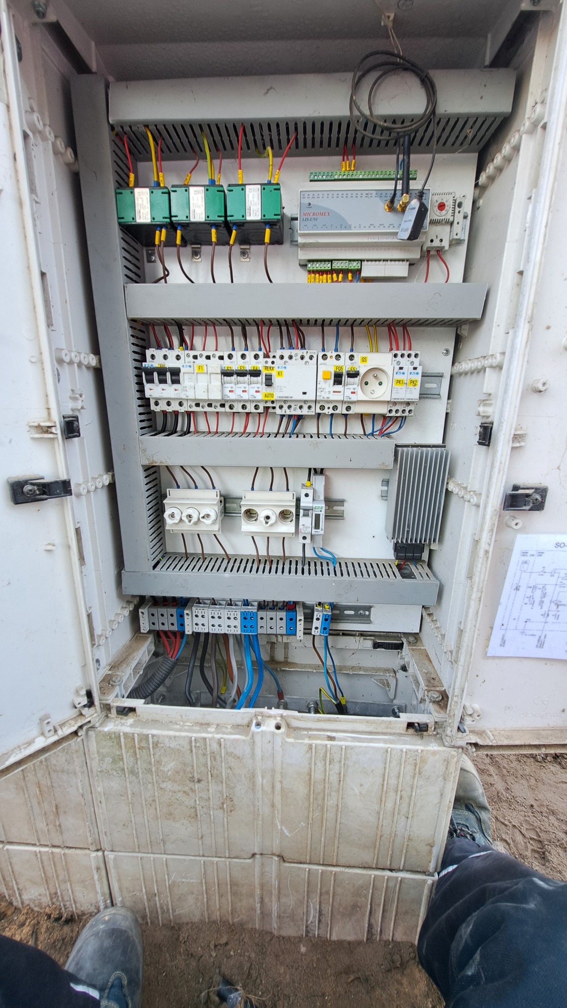 Otwarta, brudna szafa elektryczna z widocznymi bezpiecznikami, przewodami i schematem na drzwiach, sfotografowana z góry z widocznymi butami osoby stojącej przed nią.
