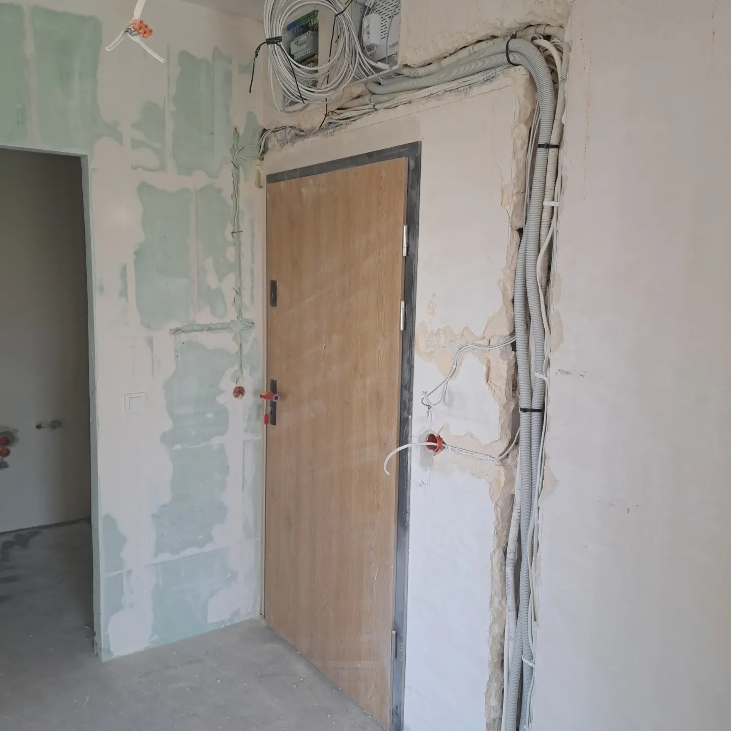 Instalacja elektryczna w trakcie montażu: kable w peszlach prowadzone wzdłuż futryny drzwi, puszki instalacyjne w ścianie, widoczna skrzynka rozdzielcza i nieotynkowane ściany w surowym stanie.
