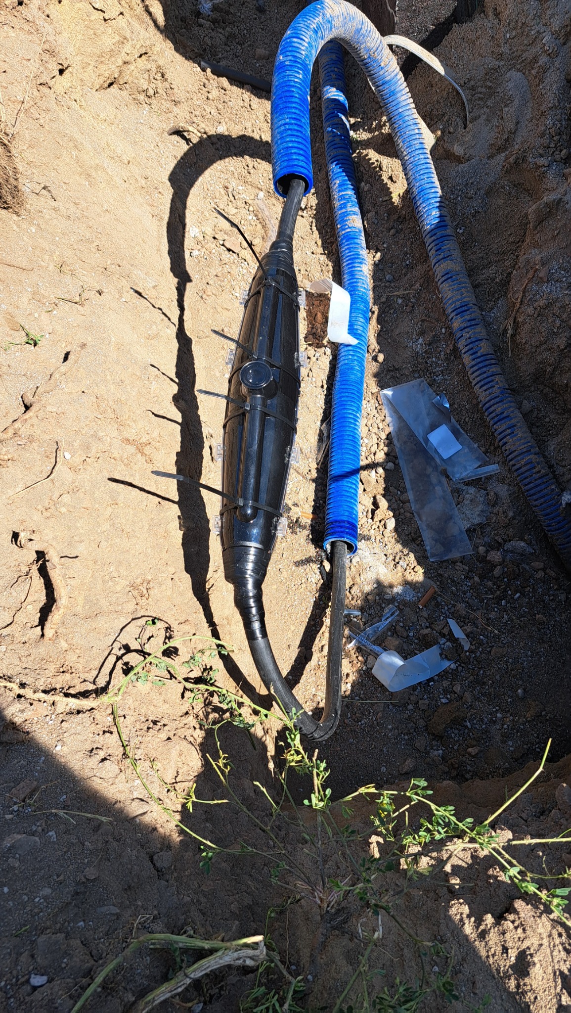 Złącze kablowe w ziemi, zabezpieczone czarną obudową i opaskami zaciskowymi, obok niebieskiego karbowanego peszla, w wykopie z piasku.