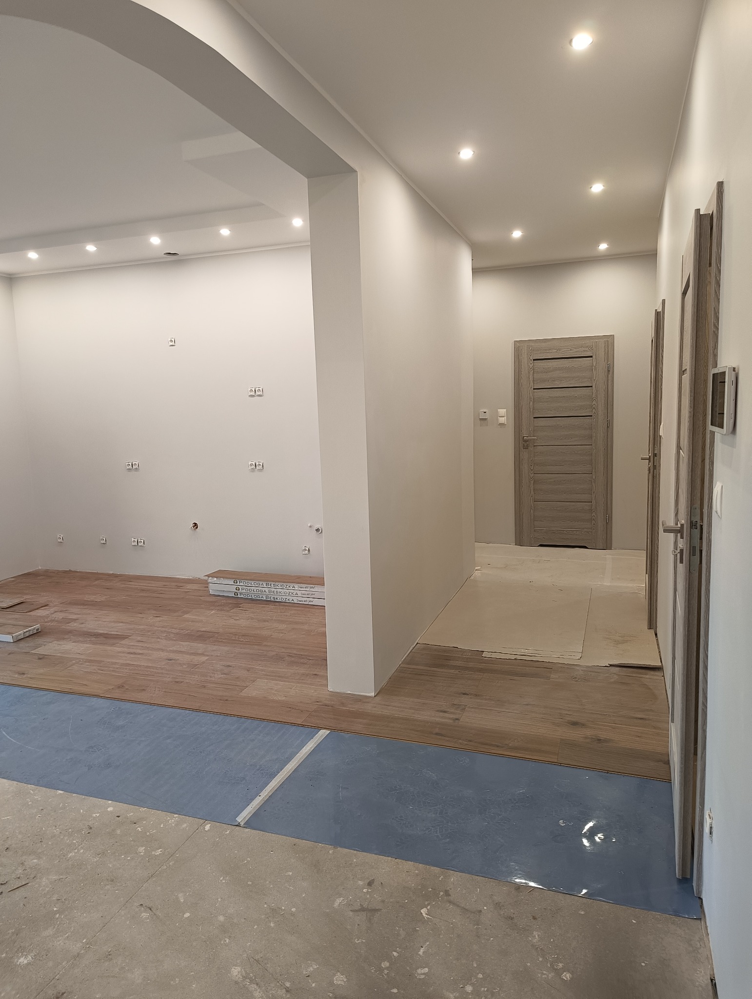 Układanie paneli podłogowych w jasnym wnętrzu w Deszcznie, widoczne puszki elektryczne, przygotowane podłoże i drzwi wewnętrzne w odcieniach szarości.