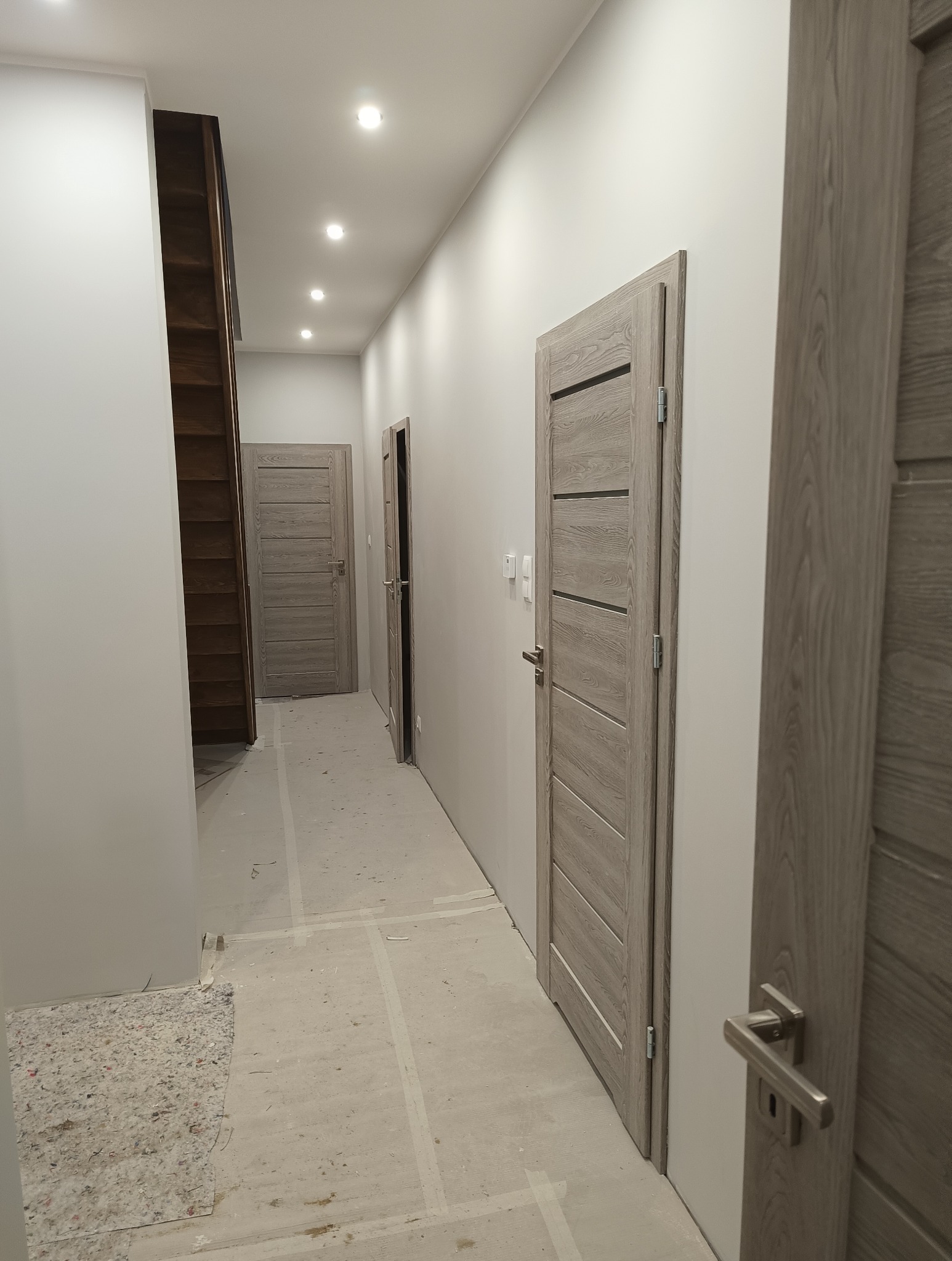 Jasny korytarz po remoncie z trzema drewnianymi drzwiami o nowoczesnym designie i punktowym oświetleniem sufitowym. Widoczne ślady prac wykończeniowych.