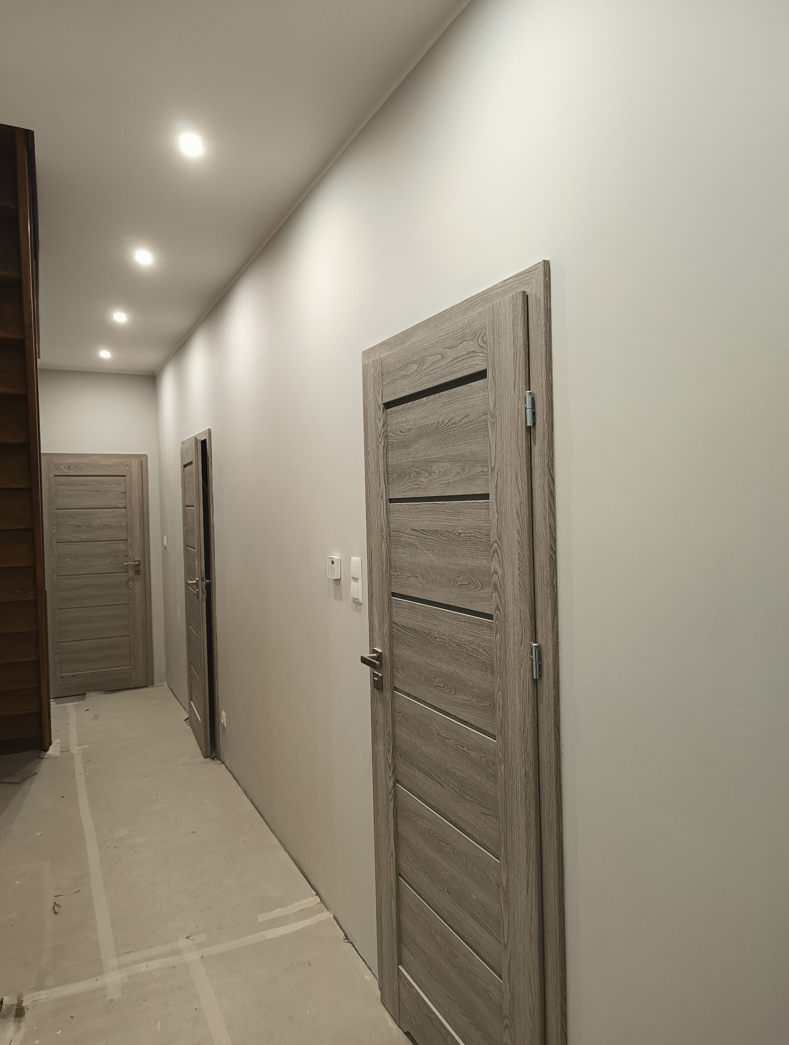 Korytarz po remoncie: szare ściany, drewniane drzwi z poziomymi zdobieniami, sufit z oświetleniem punktowym. Widoczne ślady po taśmie malarskiej na podłodze.