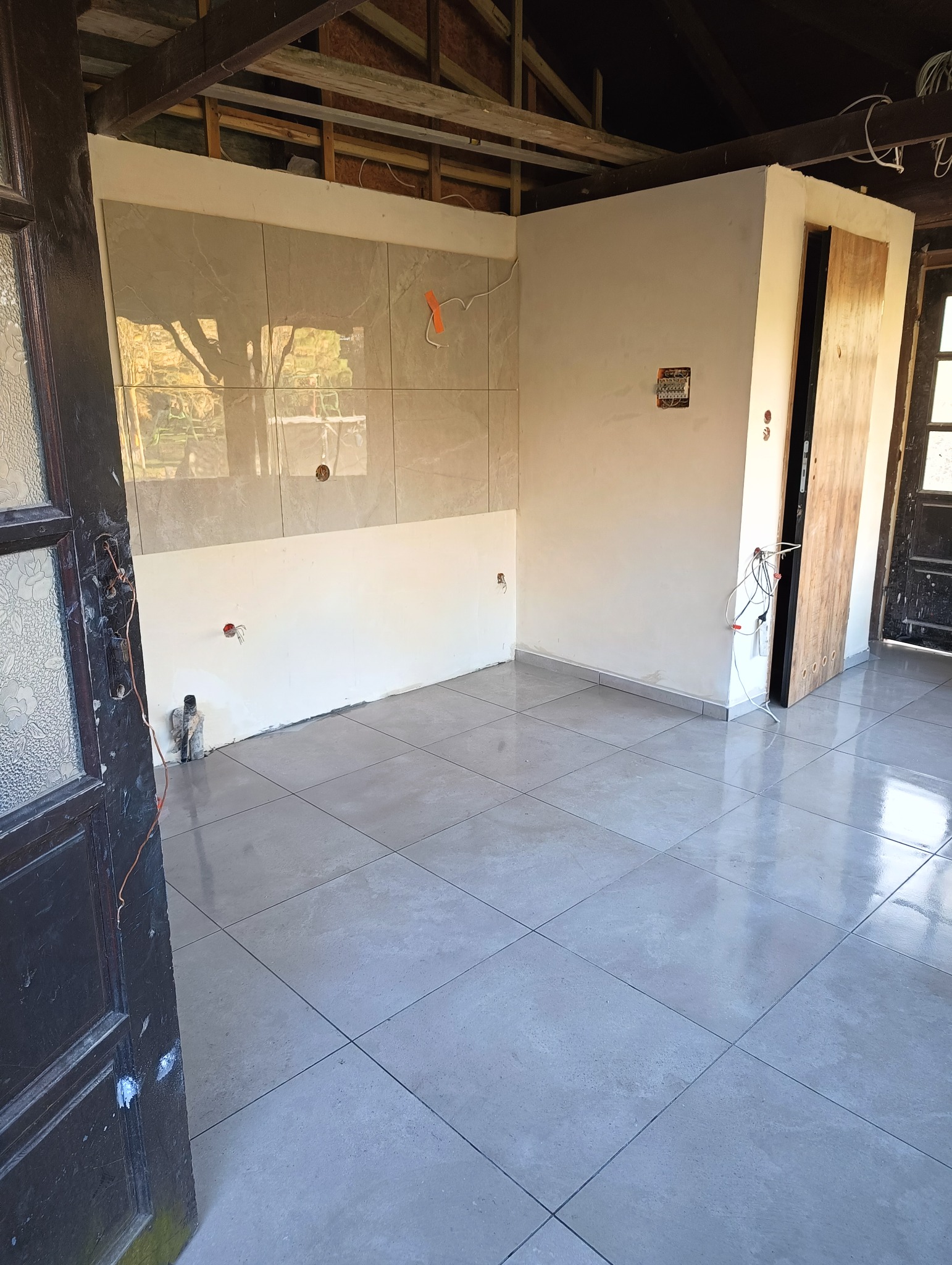 Wykończona łazienka z szarymi płytkami na podłodze i częściowo na ścianach. Widoczne przygotowane instalacje elektryczne i hydrauliczne. Drzwi wejściowe w lewym dolnym rogu.