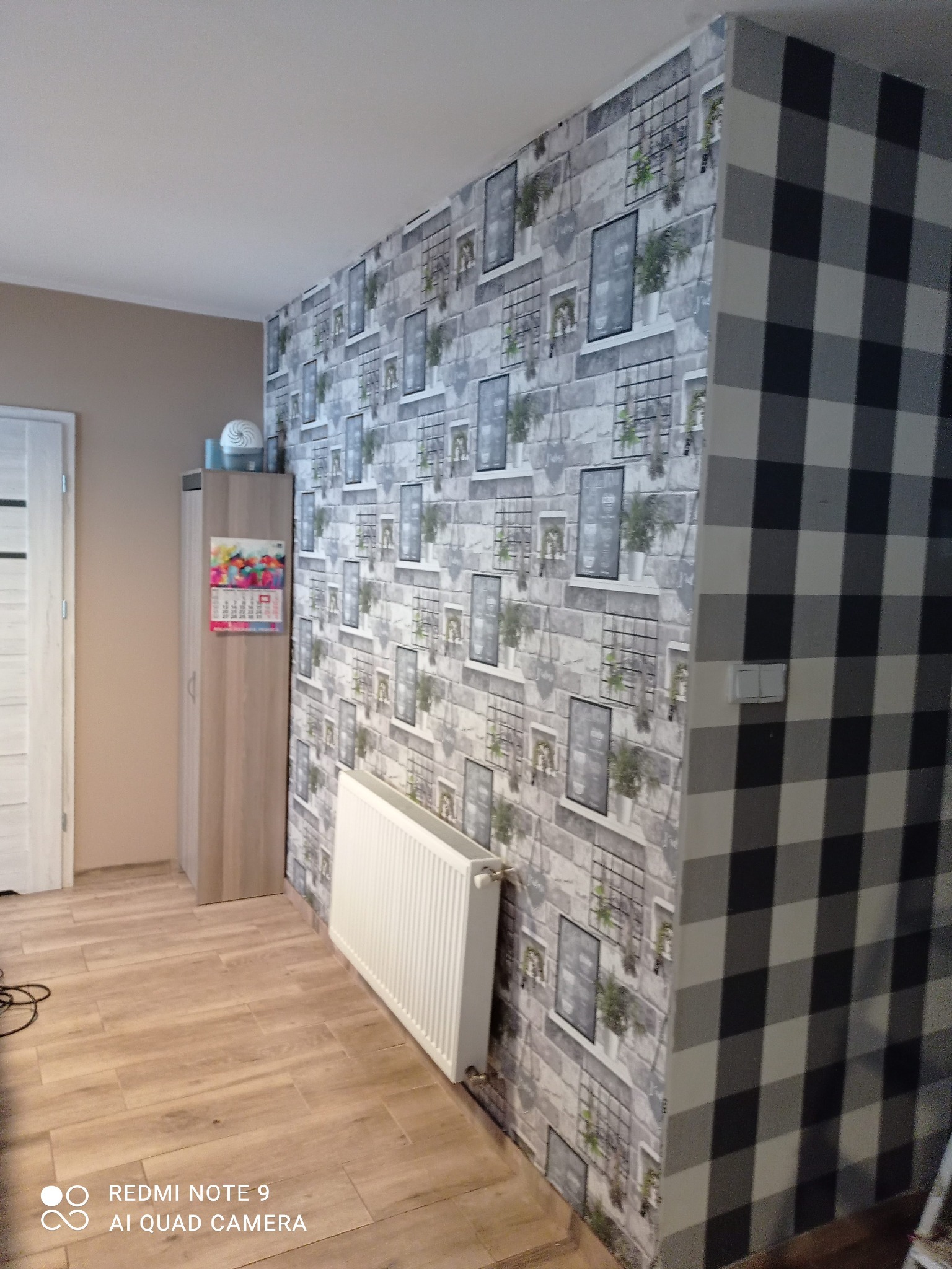 Pokój po remoncie: drewnopodobna podłoga, grzejnik, szafa z kalendarzem i ściany z tapetą w motywy cegieł i kratki. Jedna ściana w czarno-białą kratę.