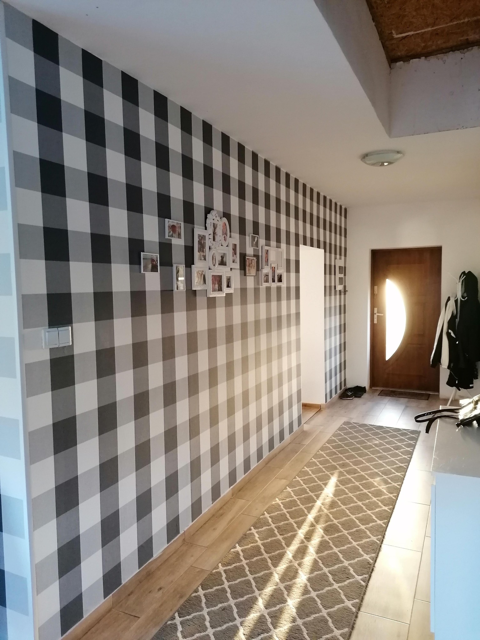 Wnętrze przedpokoju z tapetą w szaro-białą kratę. Na ścianie galeria ramek ze zdjęciami. Na podłodze dywanik w geometryczny wzór. Drzwi wejściowe z owalnym przeszkleniem.