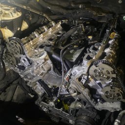 CASTROCAR SPÓŁKA Z OGRANICZONĄ ODPOWIEDZIALNOŚCIĄ - Odsłonięty silnik samochodowy w trakcie naprawy, widoczne łańcuchy rozrządu, wałki rozrządu i inne elementy wewnętrzne, oświetlenie warsztatowe.