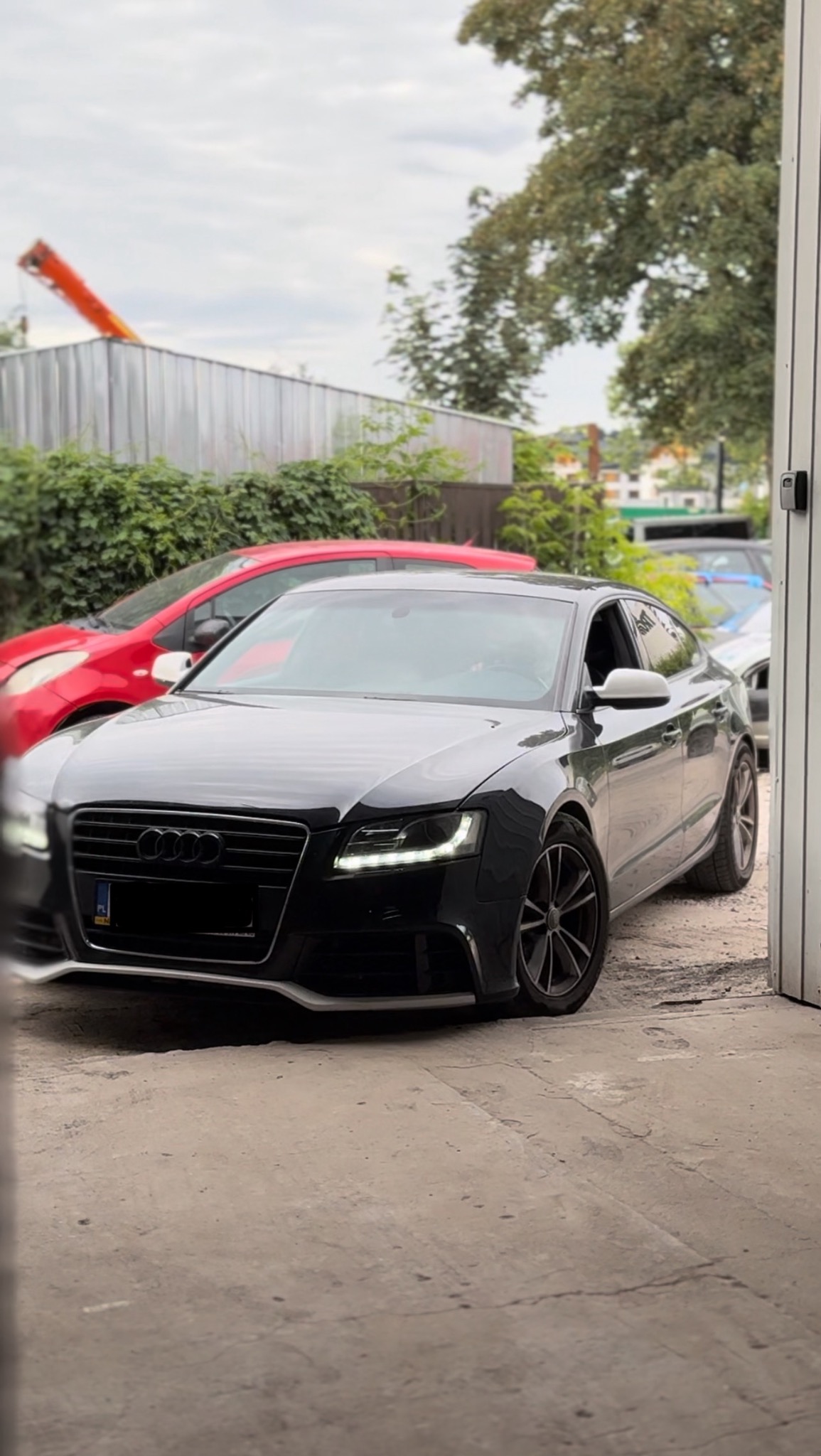 Czarny samochód Audi z włączonymi światłami LED do jazdy dziennej, zaparkowany przed garażem, z widocznymi innymi pojazdami w tle.