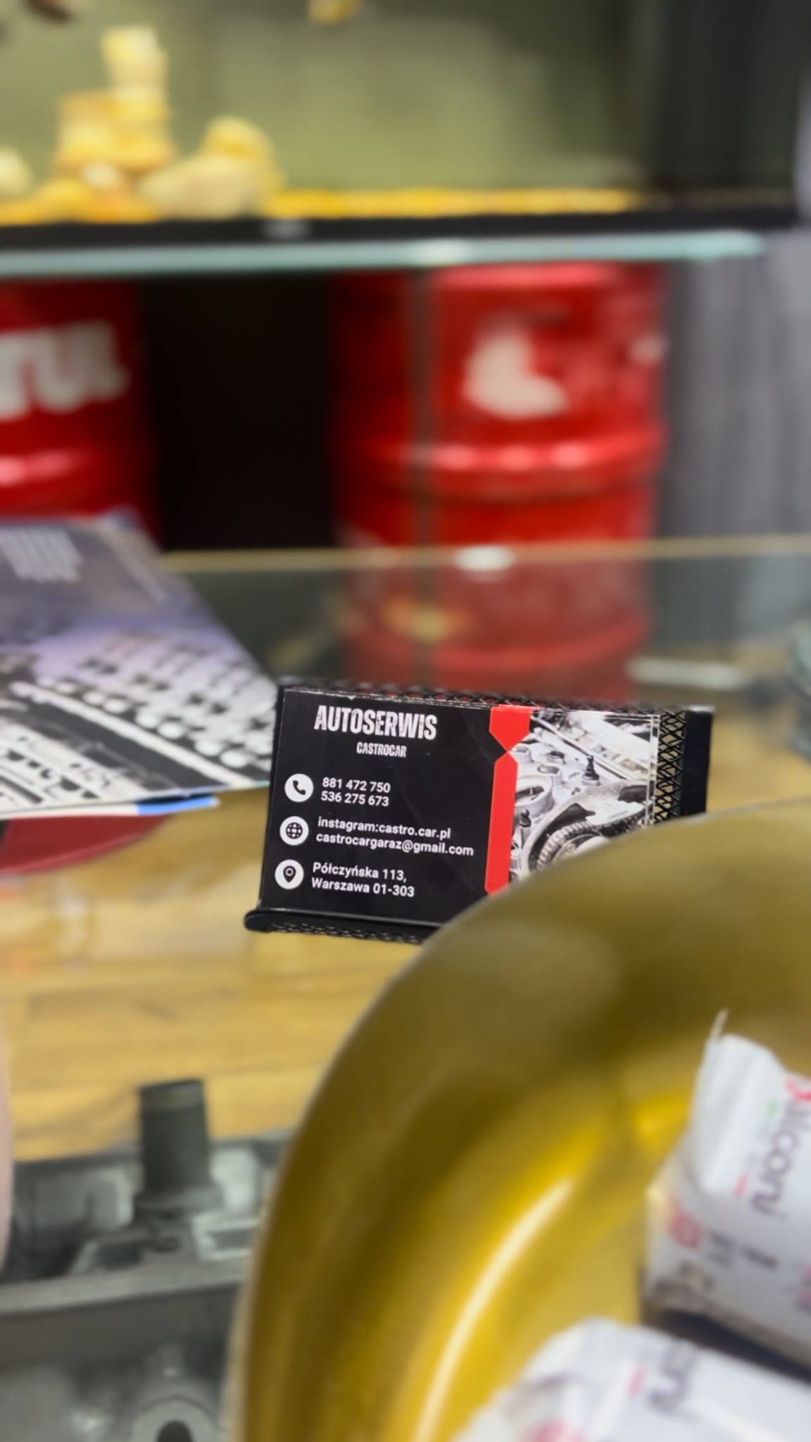 Wizytówka AUTOSERWIS Castrocar z numerami telefonów, adresem i Instagramem na tle części samochodowych i czerwonych beczek z olejem.