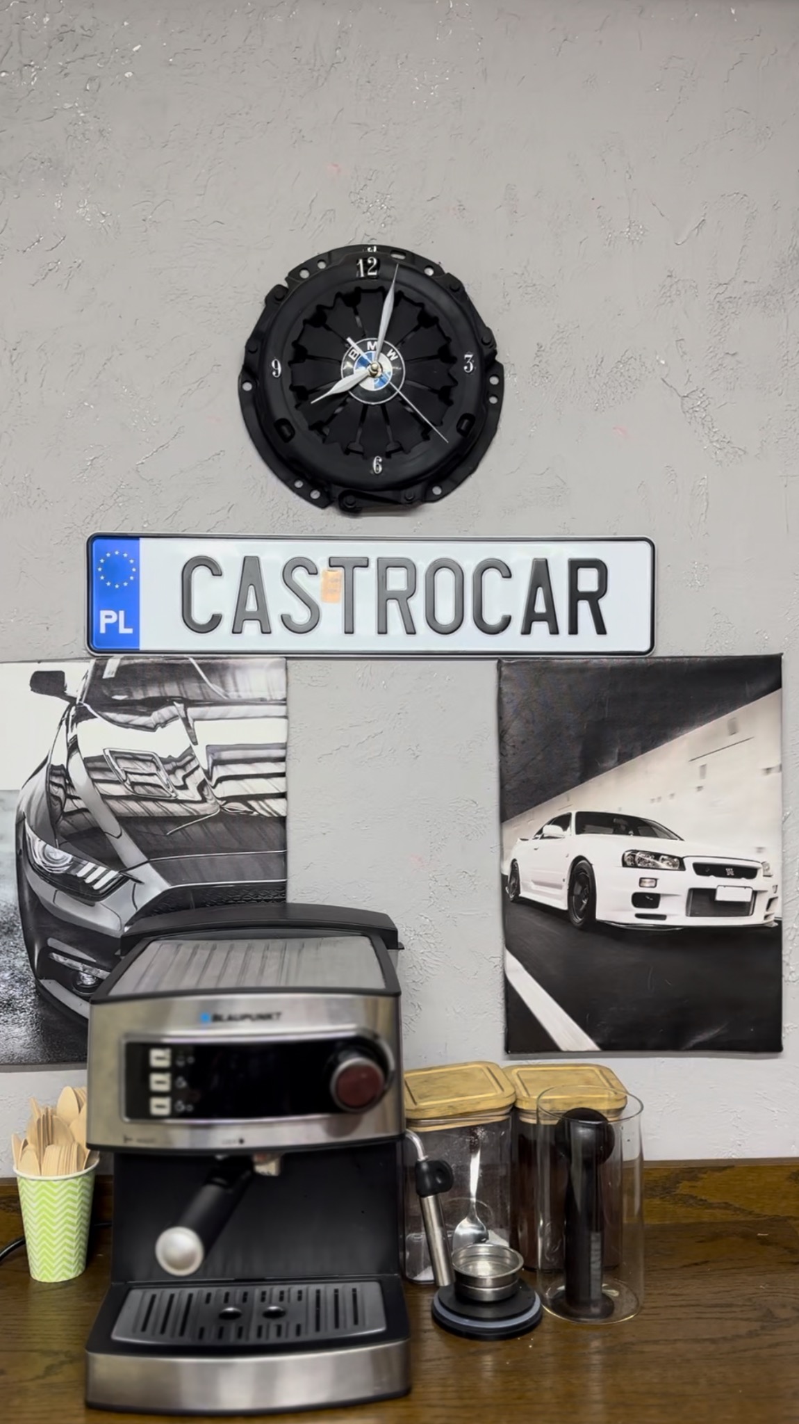 Warsztatowy wystrój wnętrza: zegar z tarczy sprzęgła BMW, tablica rejestracyjna 'CASTROCAR', obrazy z samochodami sportowymi, ekspres do kawy.