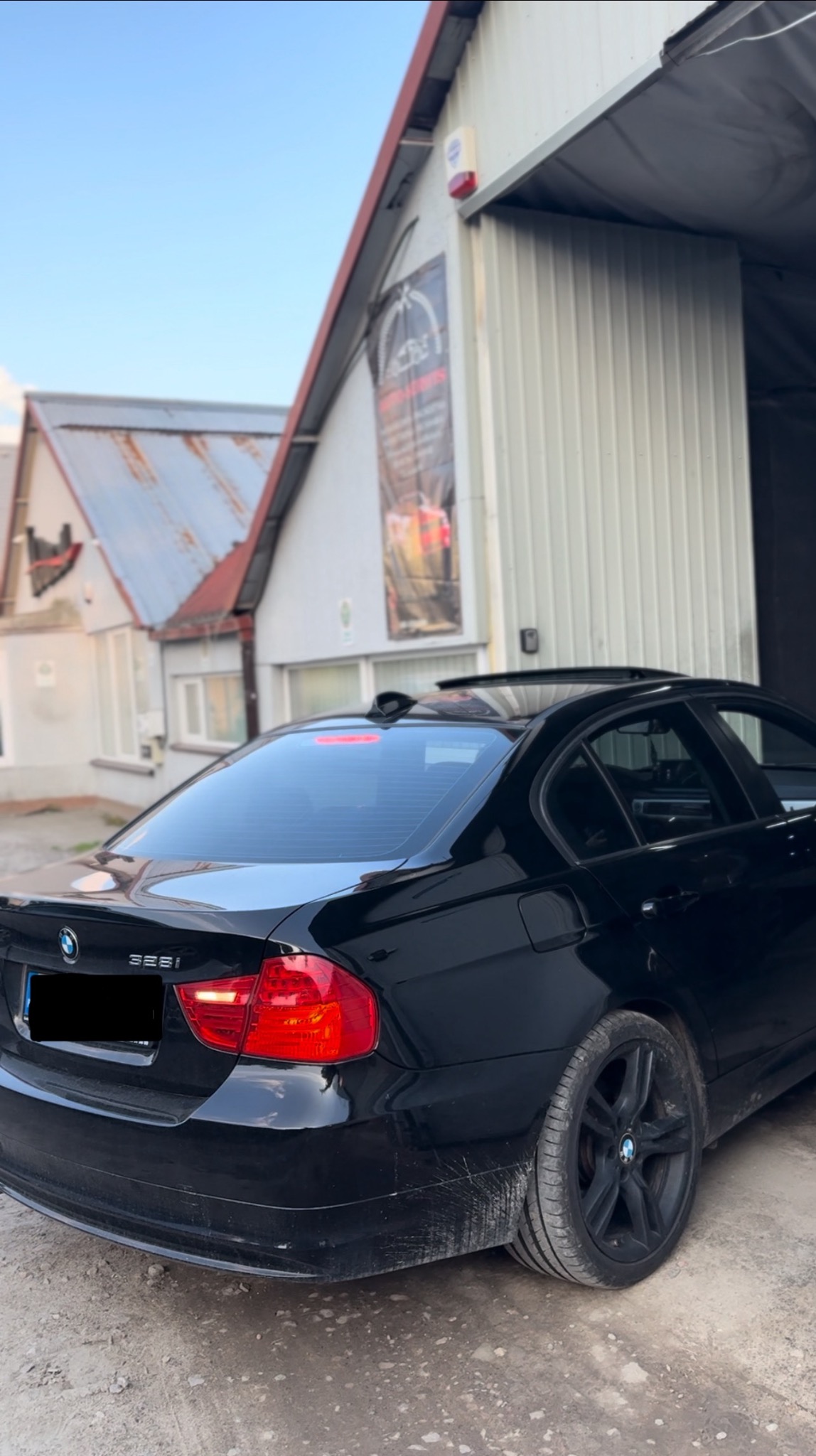 Czarny samochód BMW 325i zaparkowany przed budynkiem warsztatu z metalową elewacją i szyldem reklamowym, widok z tyłu.