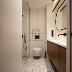 Klaudia Gołębiewska - Nowoczesna łazienka z kabiną prysznicową typu walk-in, umywalką z drewnianą szafką, okrągłym lustrem z podświetleniem LED i podłogą z lastryko w odcieniach szarości i bieli.