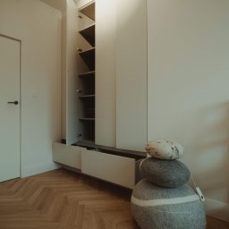 Klaudia Gołębiewska - Minimalistyczne wnętrze z zabudową meblową w odcieniach bieli, drewnianą podłogą w jodełkę i dekoracyjnymi poduszkami ułożonymi w stos. Otwarta szafka z półkami ukazuje przechowywane przedmioty.