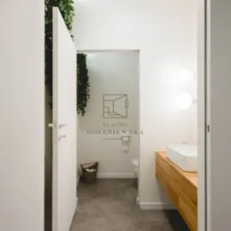 Nowoczesna łazienka z betonową podłogą, wiszącą zielenią i drewnianą szafką pod umywalką, widziana zza uchylonych drzwi. Logo projektanta wnętrz na ścianie.