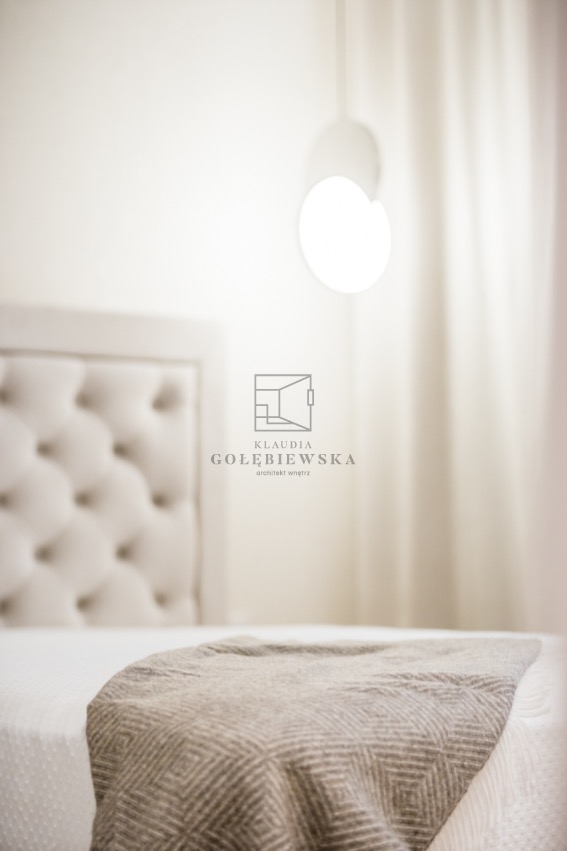 Delikatne ujęcie sypialni z pikowanym zagłówkiem łóżka, zwisającą okrągłą lampą i pledem w geometryczny wzór, przysłonięte logo architekta wnętrz.